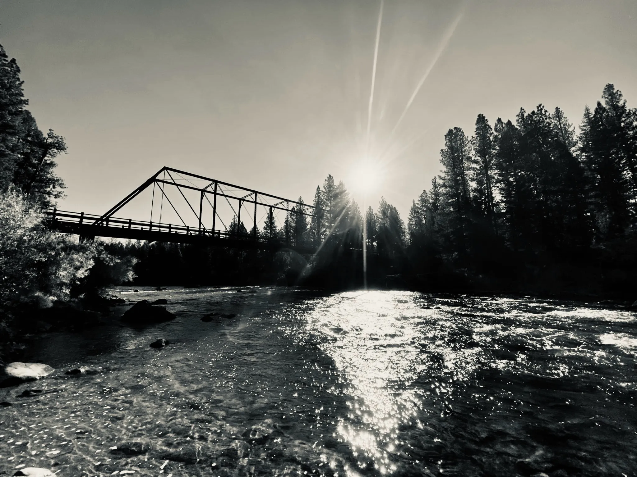 A metal truss bridge crosses a rocky river as the sun shines through trees behind it.