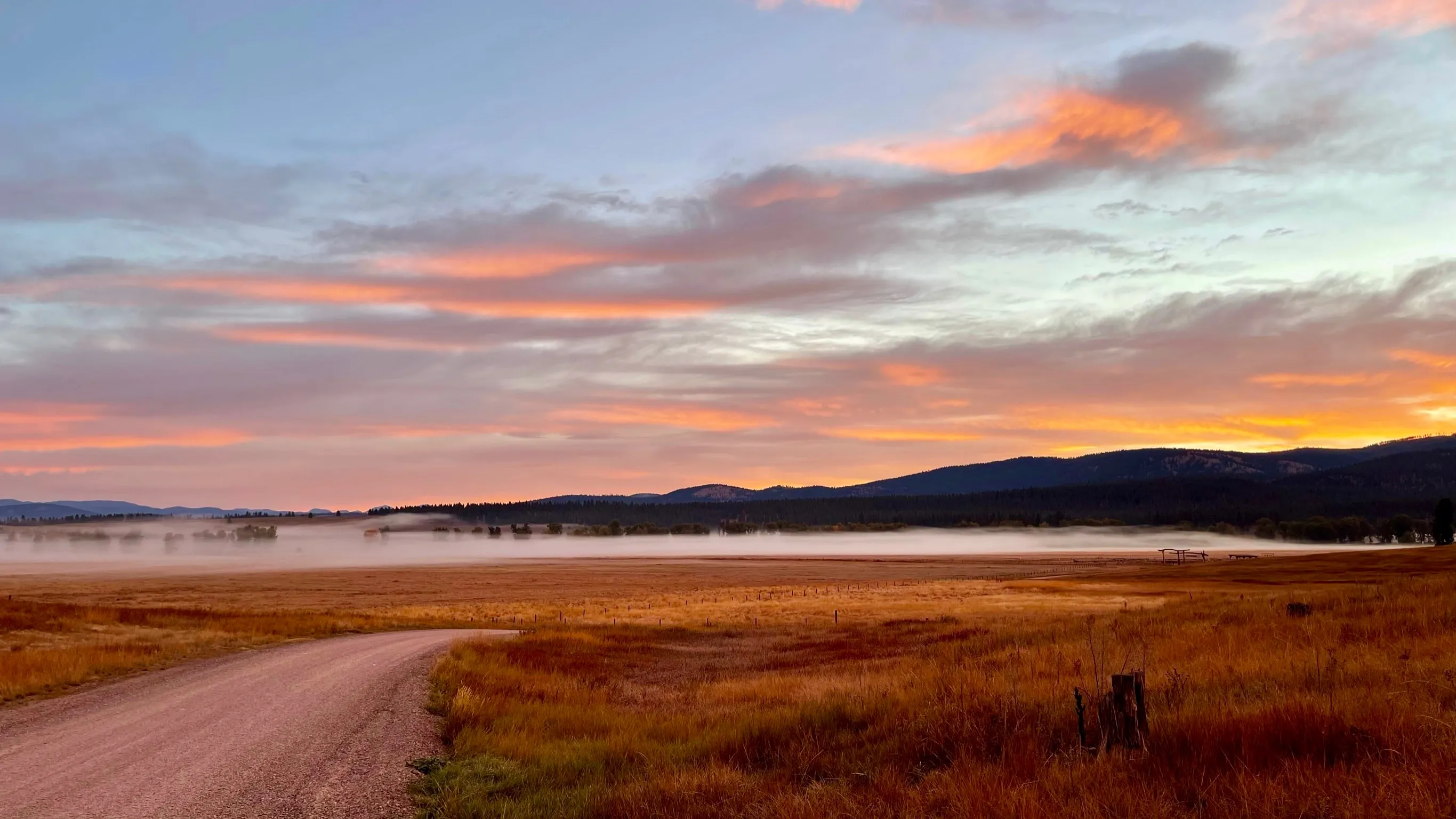 A pale gravel road curves through brown fields toward low dark hills under pink and orange clouds.