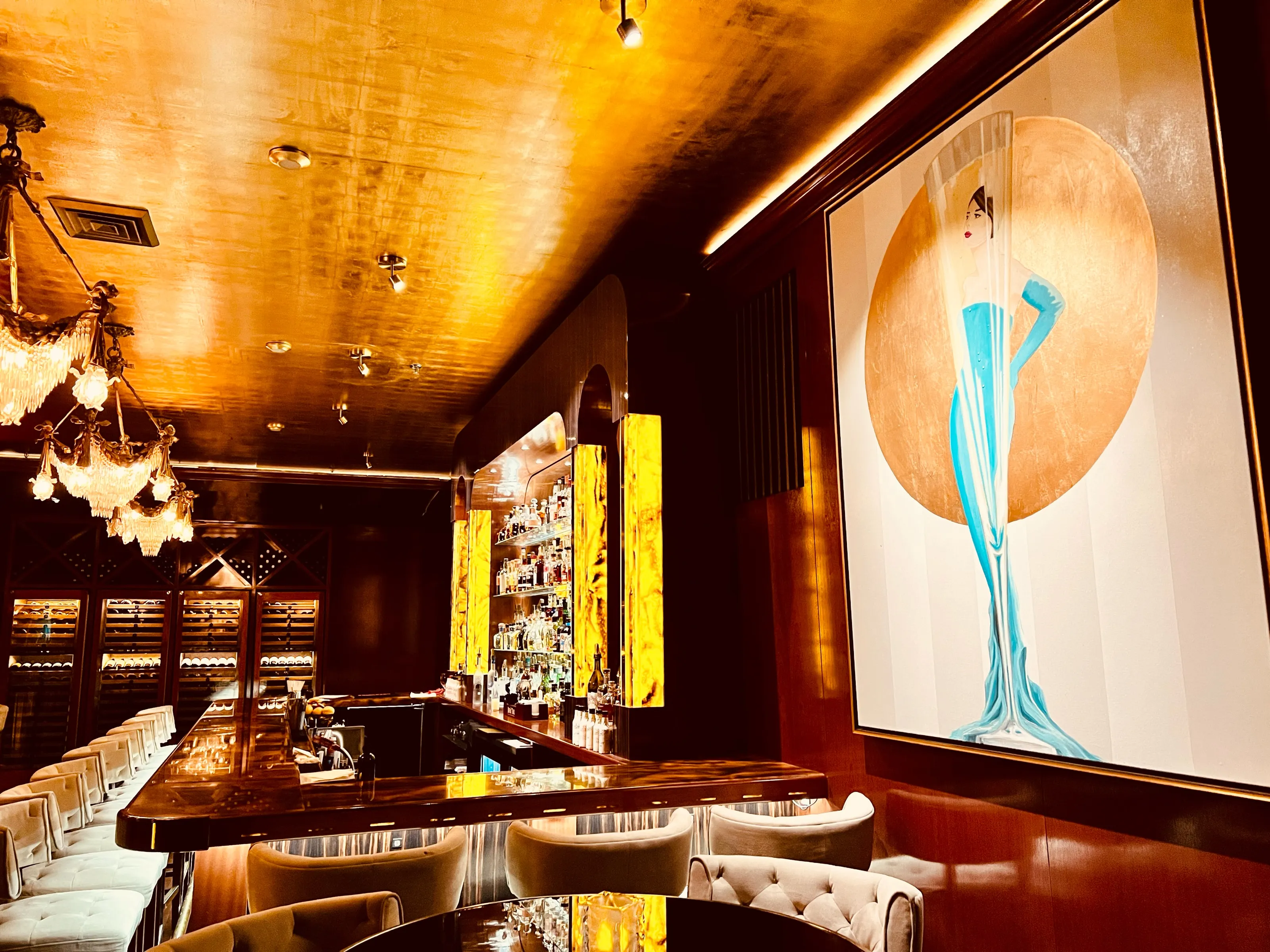 A warmly lit bar interior with chandeliers, seating, a long counter, and a large painting on the wall.