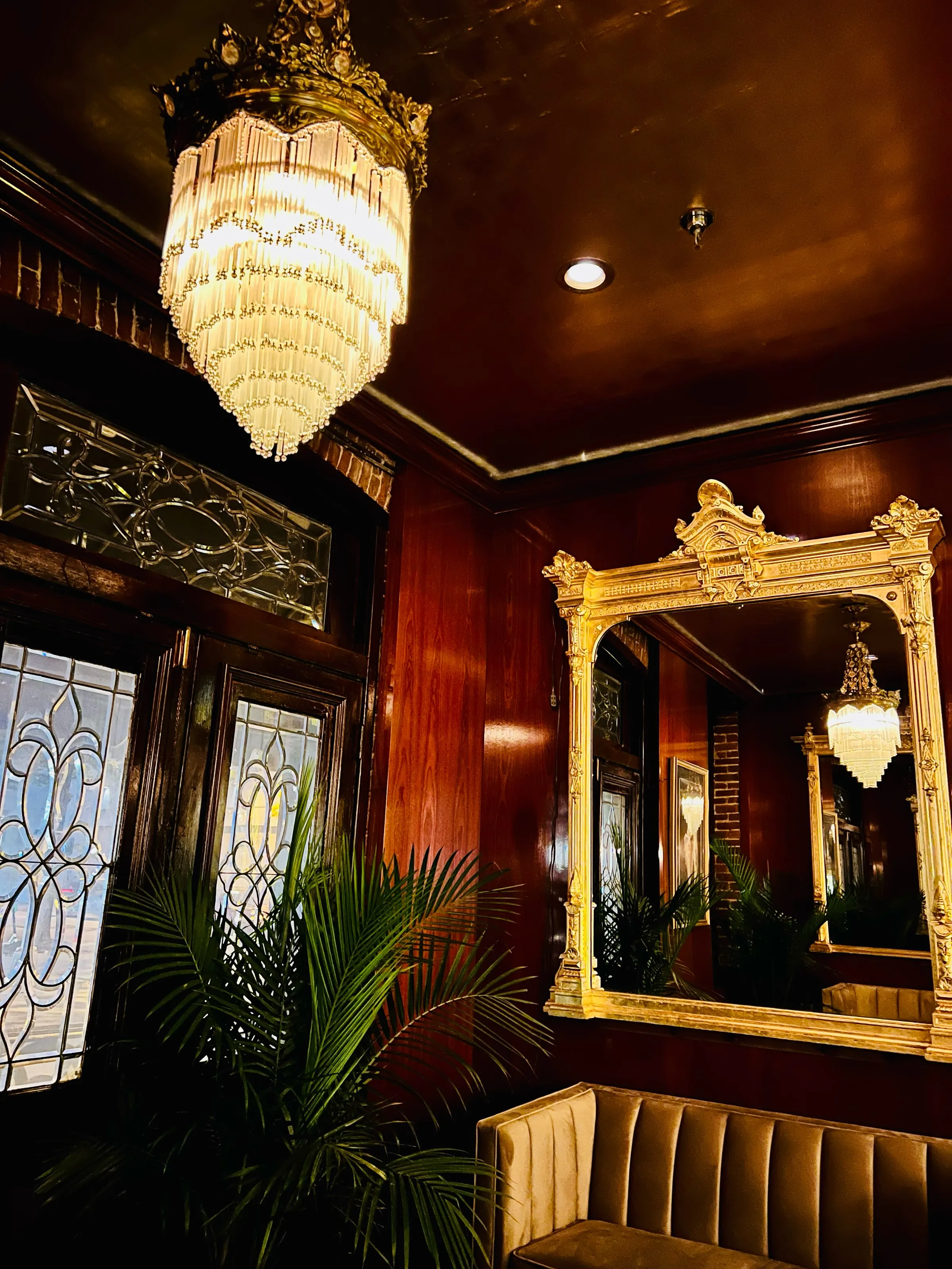 A warm wood-paneled room with a crystal chandelier, gilded mirror, stained glass, and a velvet bench.