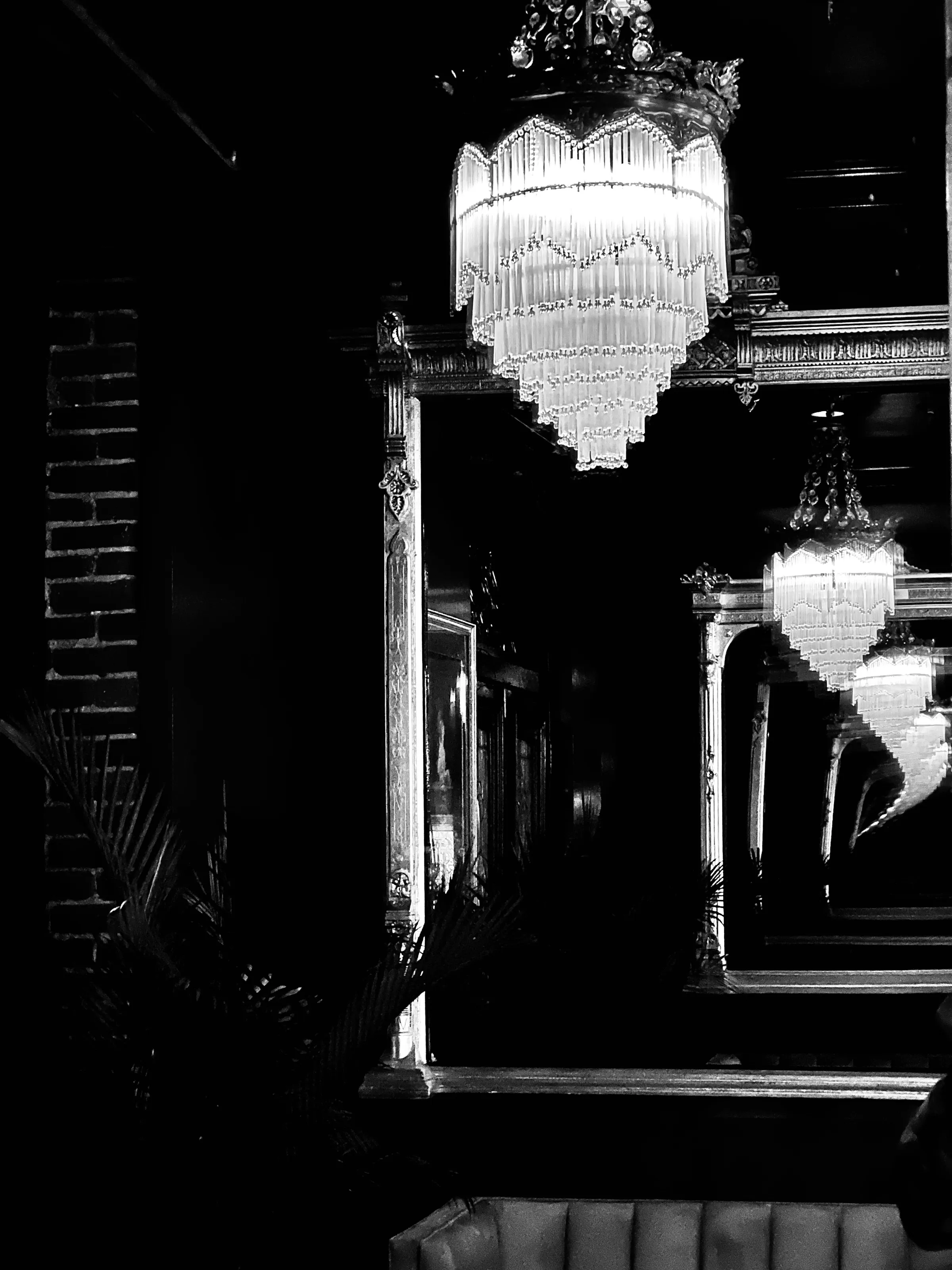 Two ornate chandeliers hang in a dark interior, with one reflected in a mirror.
