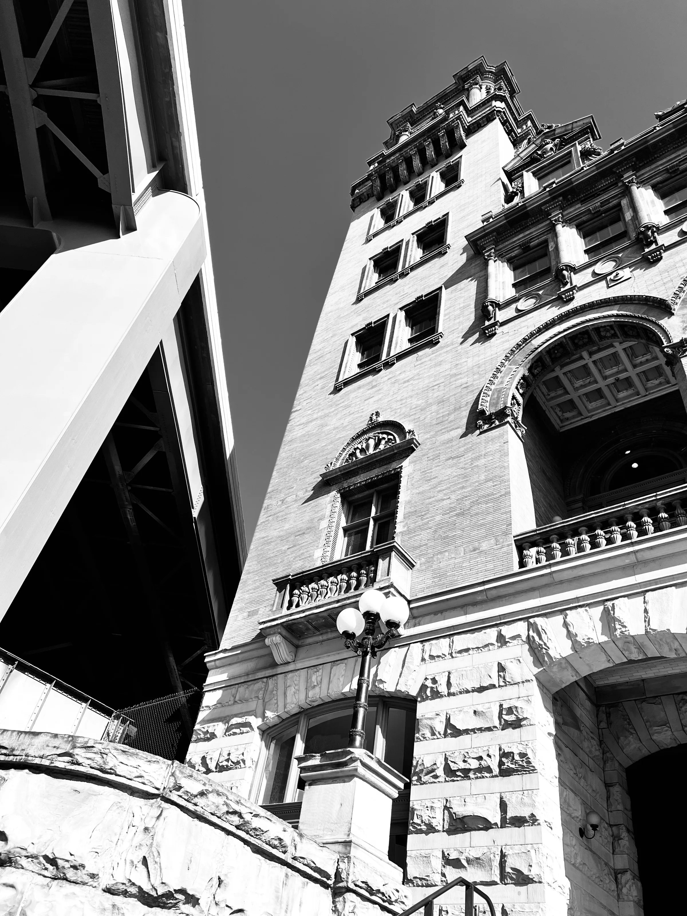 A black-and-white low-angle view of an ornate stone building next to a large bridge support.