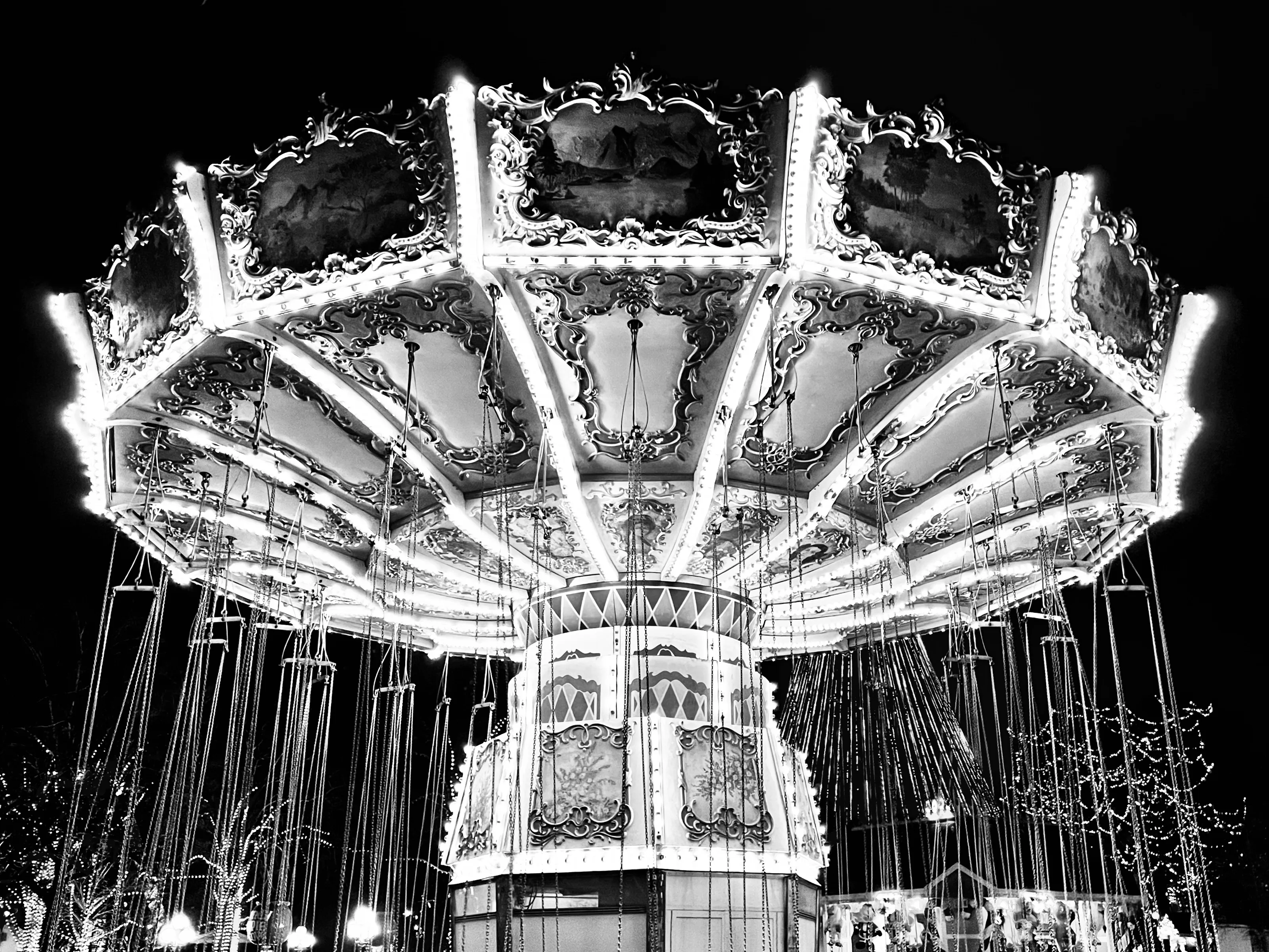Black-and-white upward view of an ornate illuminated swing carousel with empty chains hanging around its circular canopy.