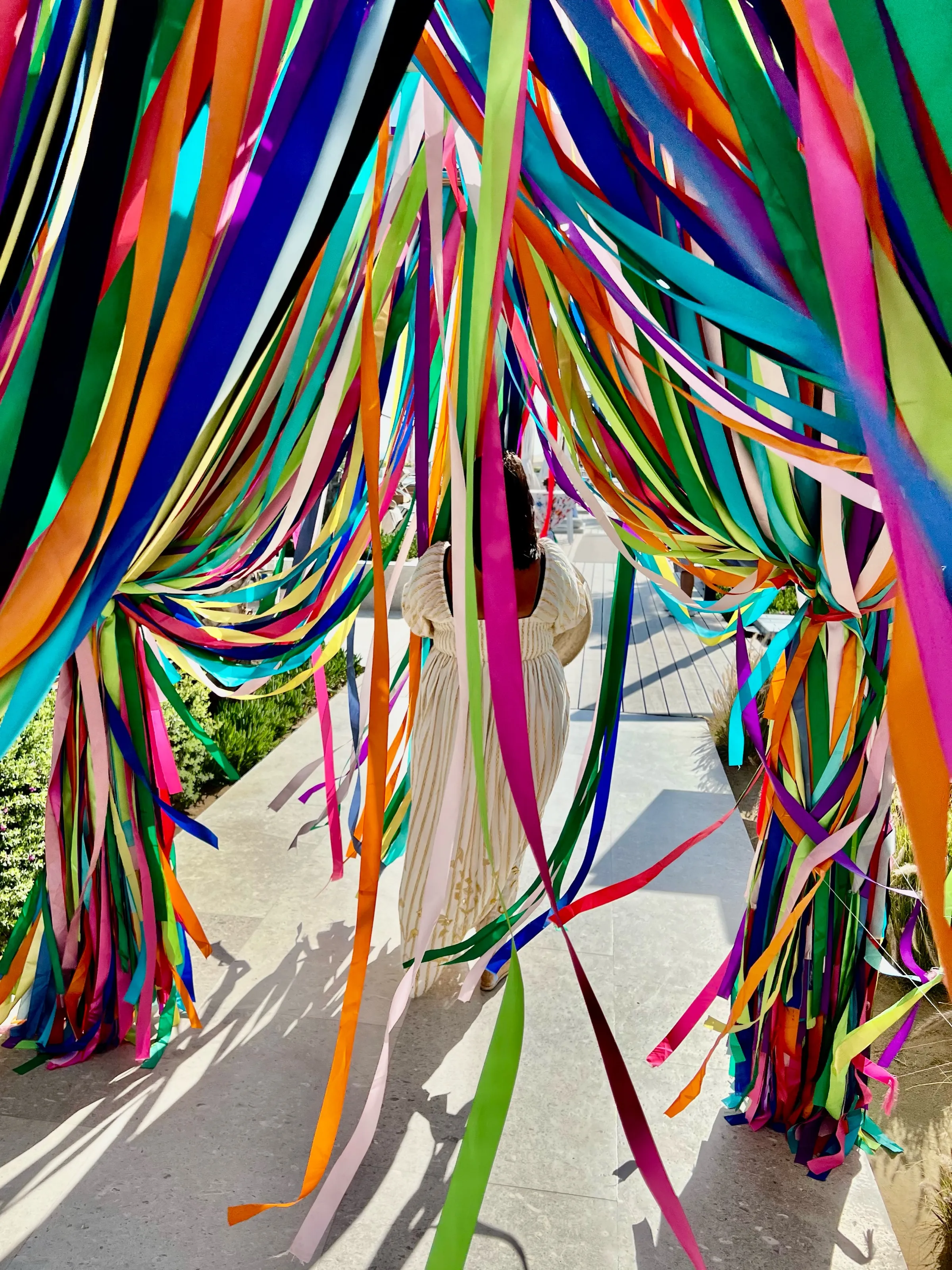 A person in a light dress walks through a passage filled with long multicolored ribbons hanging overhead.