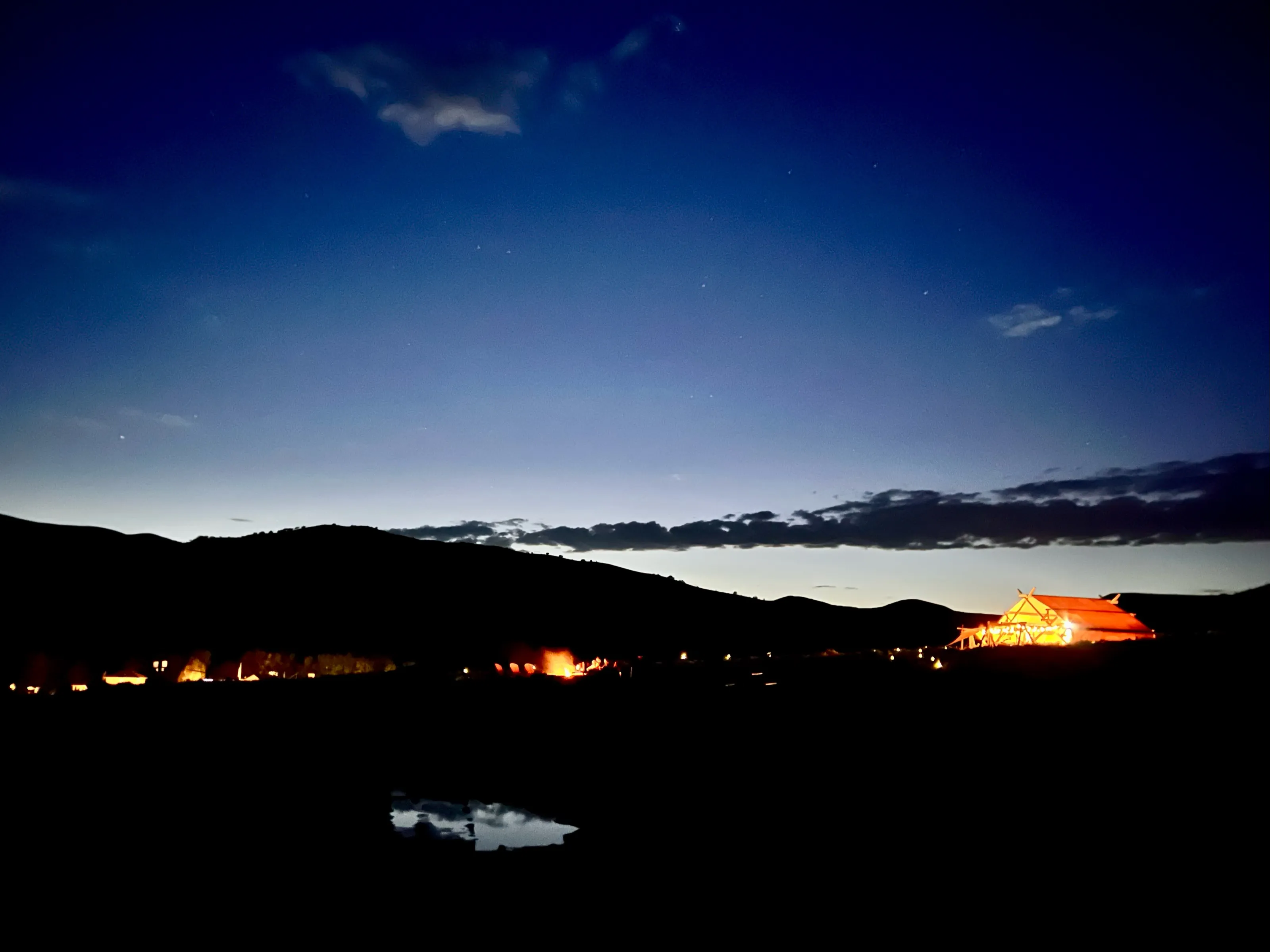 A dark mountain ridge and scattered warm lights sit below a deep blue evening sky, with a small puddle reflecting light in the foreground.