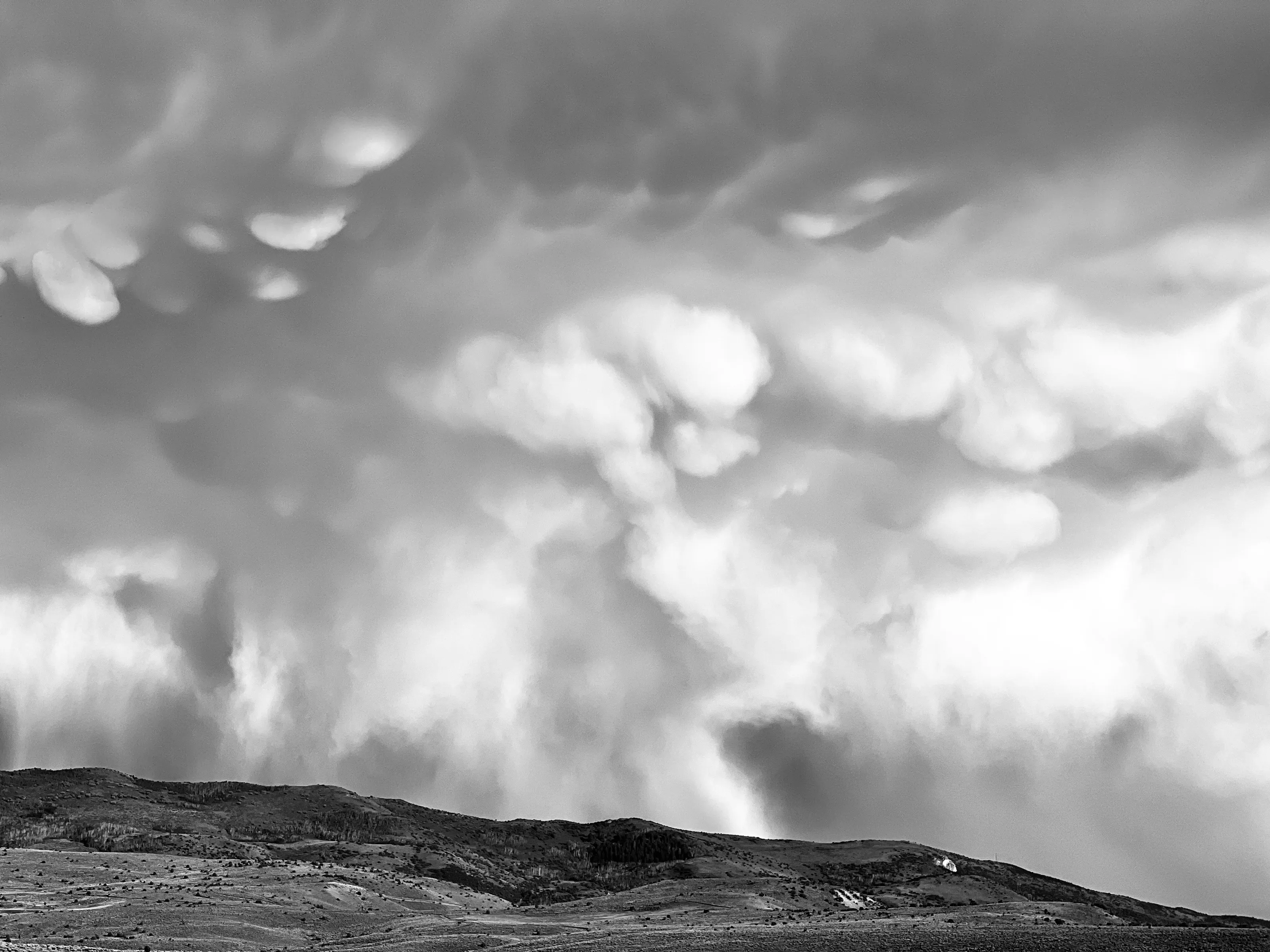 Black-and-white view of low textured hills beneath thick clouds and pale vertical curtains of rain.
