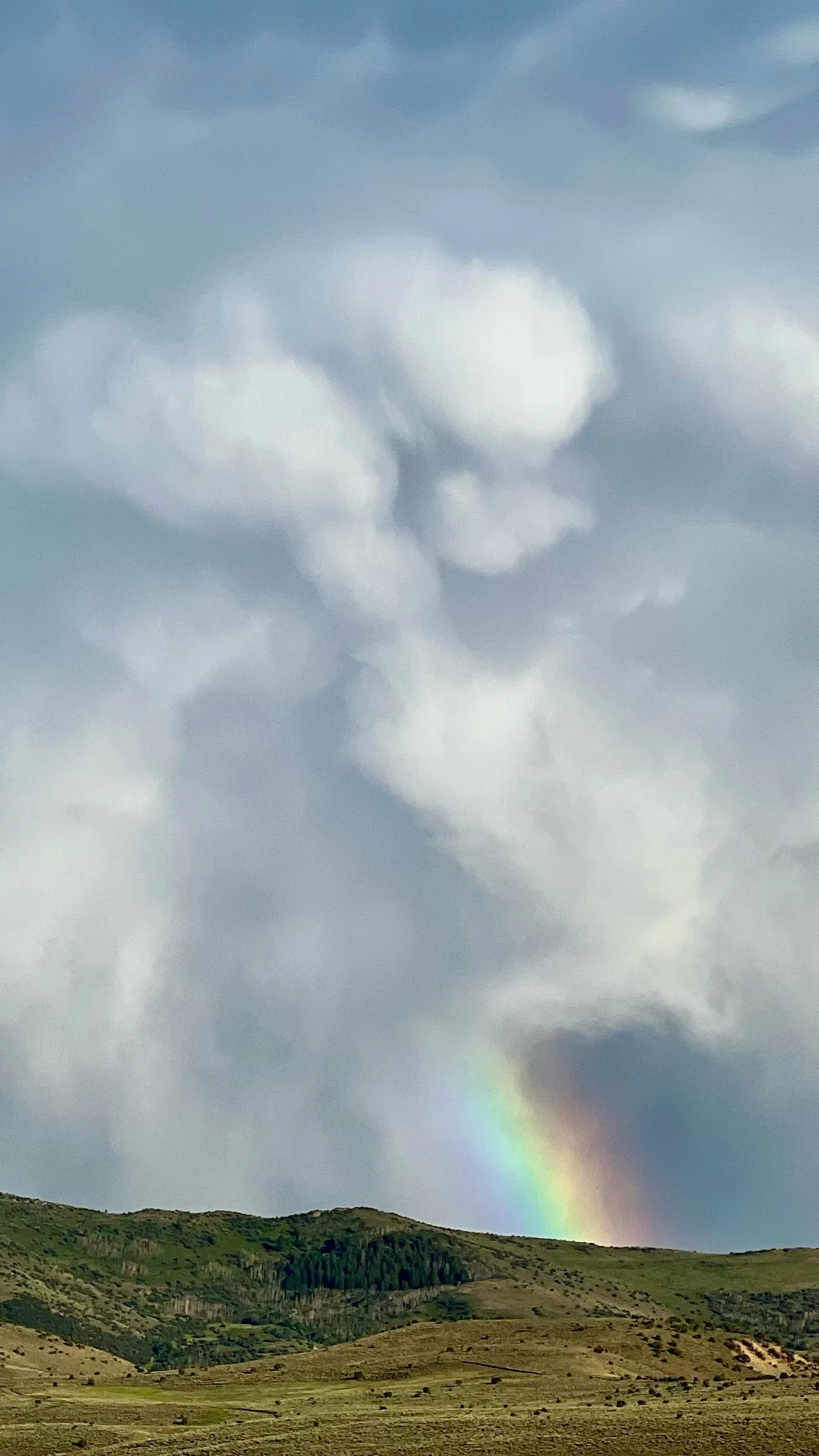 A faint rainbow beneath large pale storm clouds over green rolling hills.