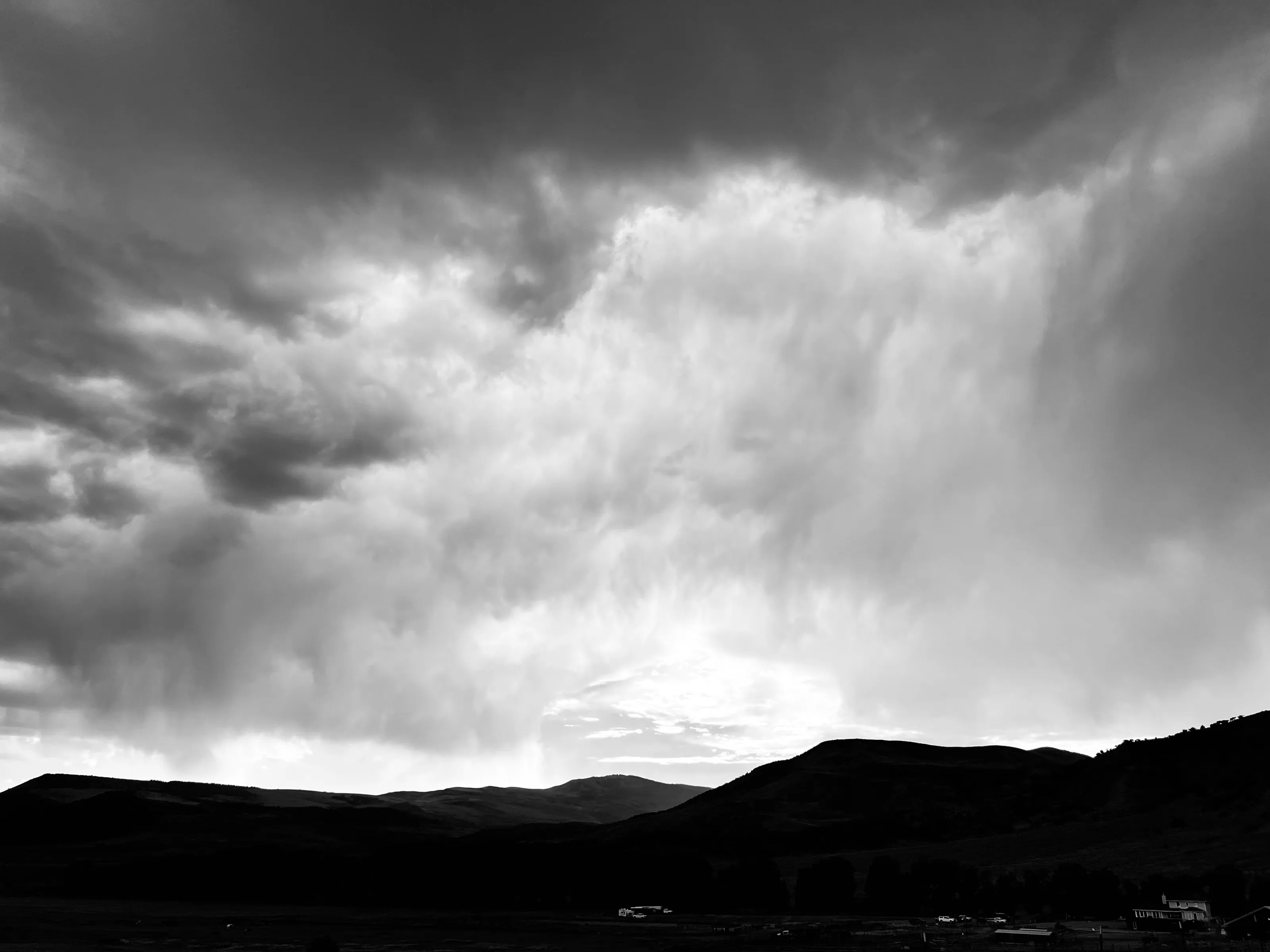 Black-and-white panorama of dark mountain ridges with pale rain shafts falling from thick clouds.