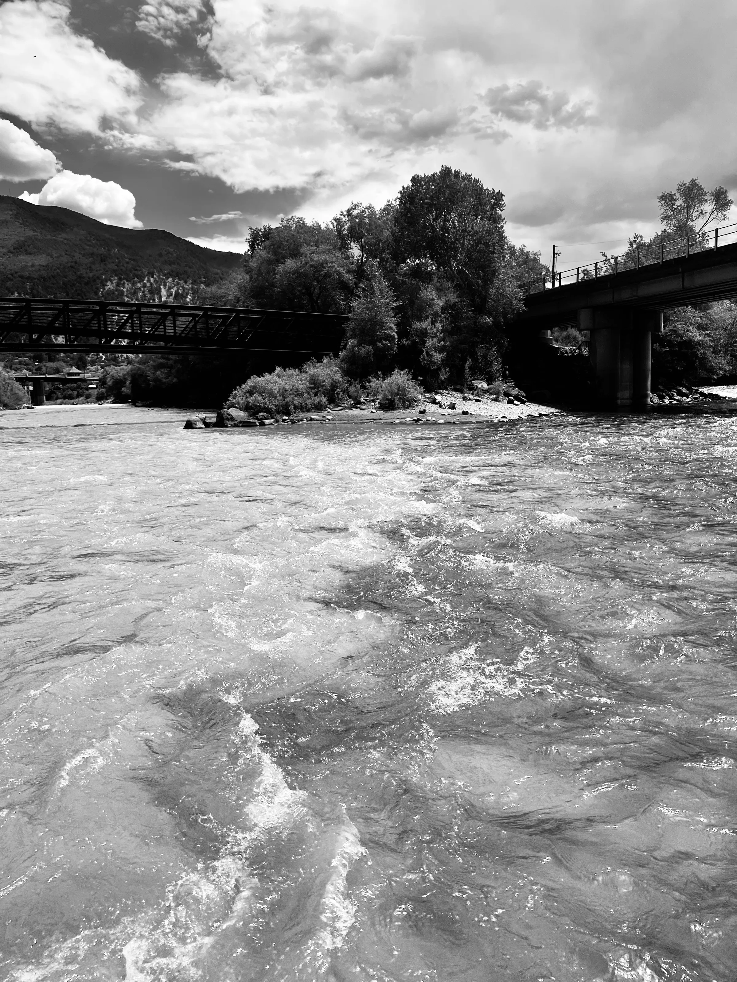 Black-and-white view of a fast river flowing between a pedestrian bridge and a road bridge, with trees and mountains behind.