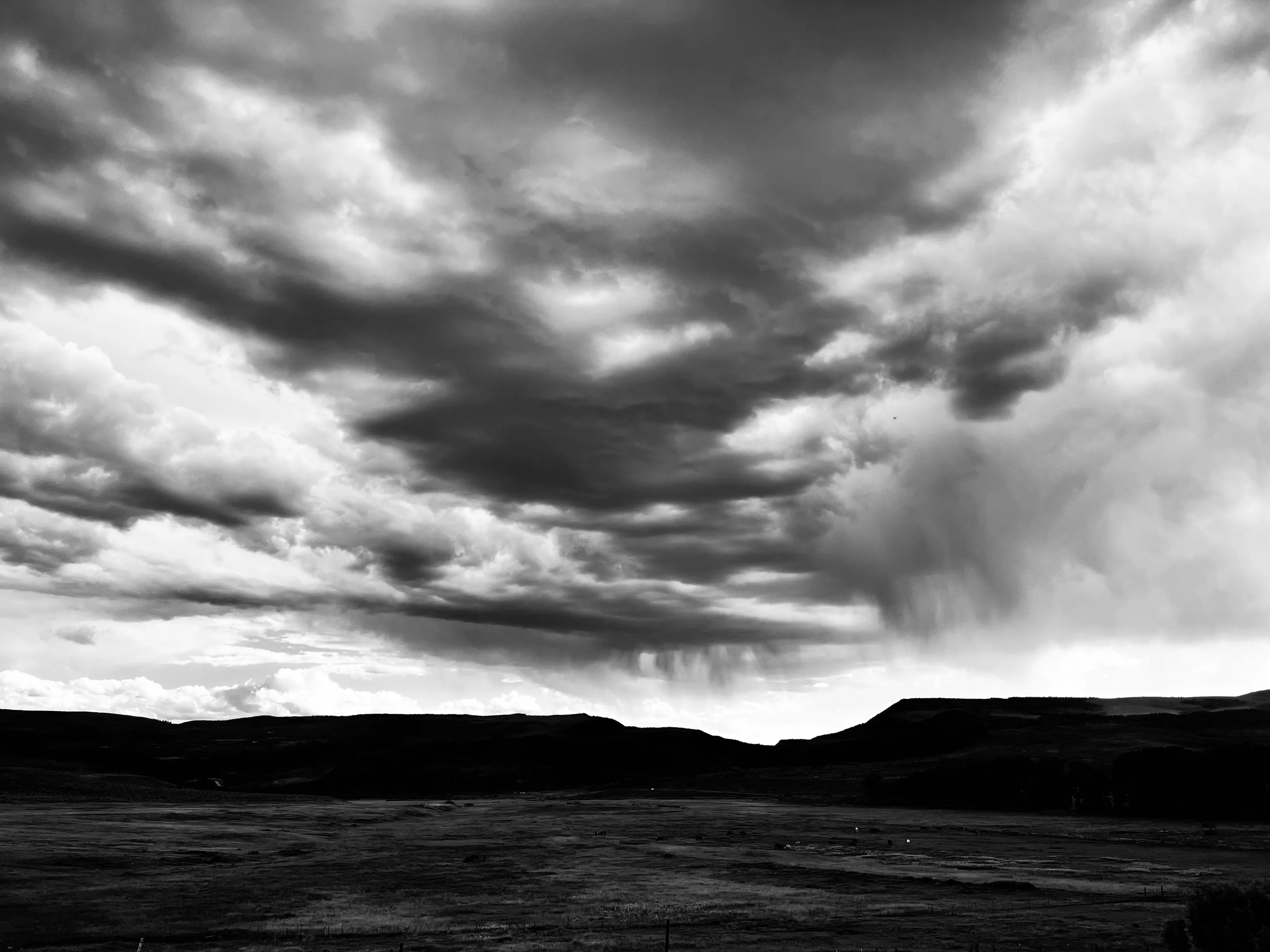 Black-and-white landscape of dark low hills under dramatic clouds and a curtain of rain.