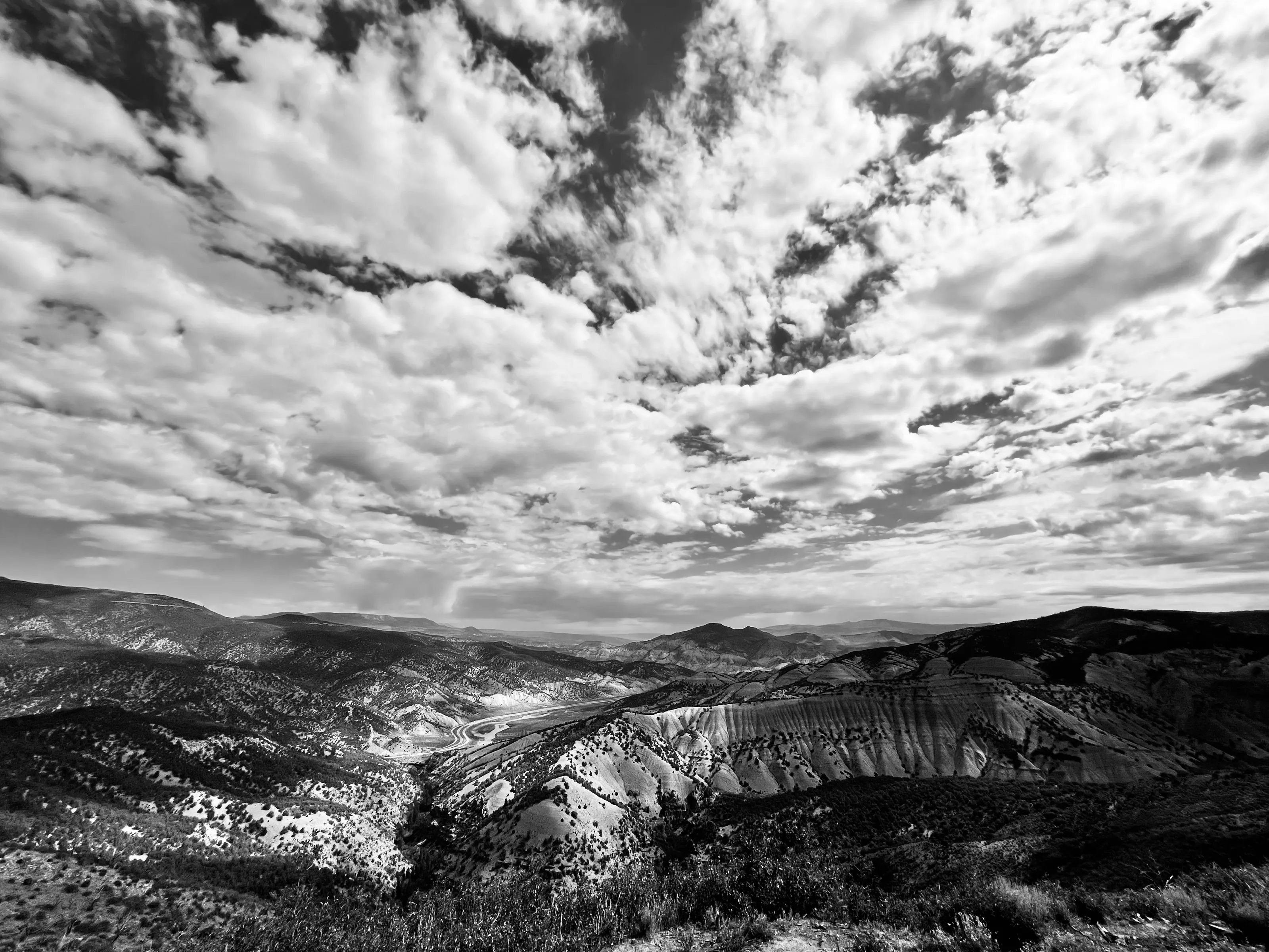 Black-and-white landscape of textured clouds over a broad valley with eroded hills and dark foreground vegetation.