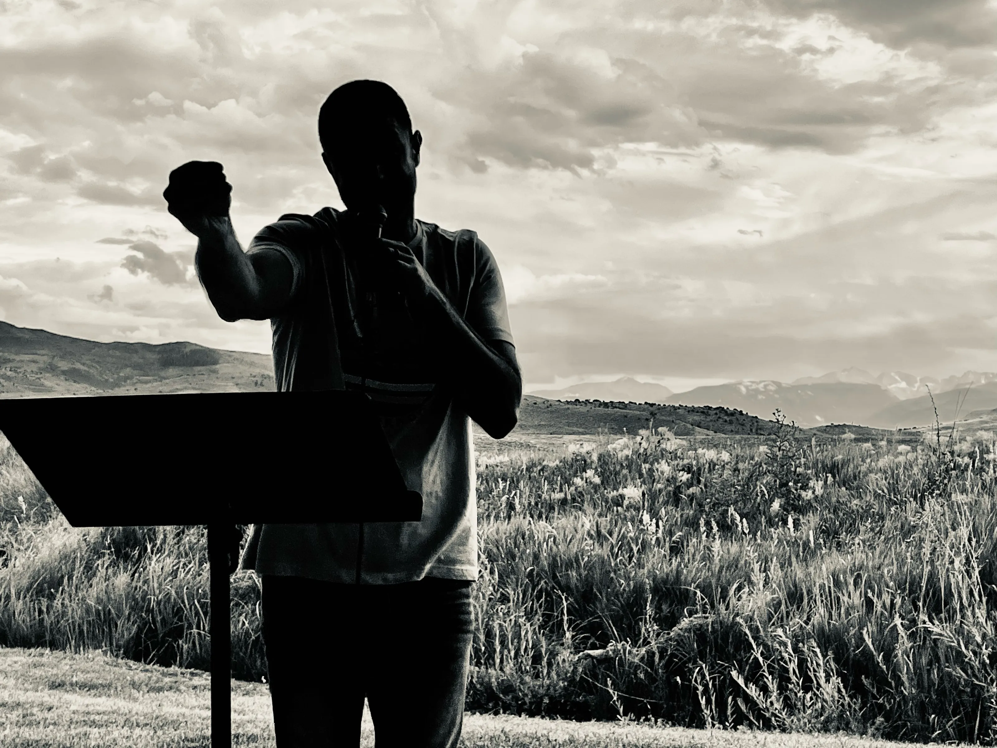Black-and-white photograph of a person in silhouette holding a microphone beside a music stand, with hills behind.