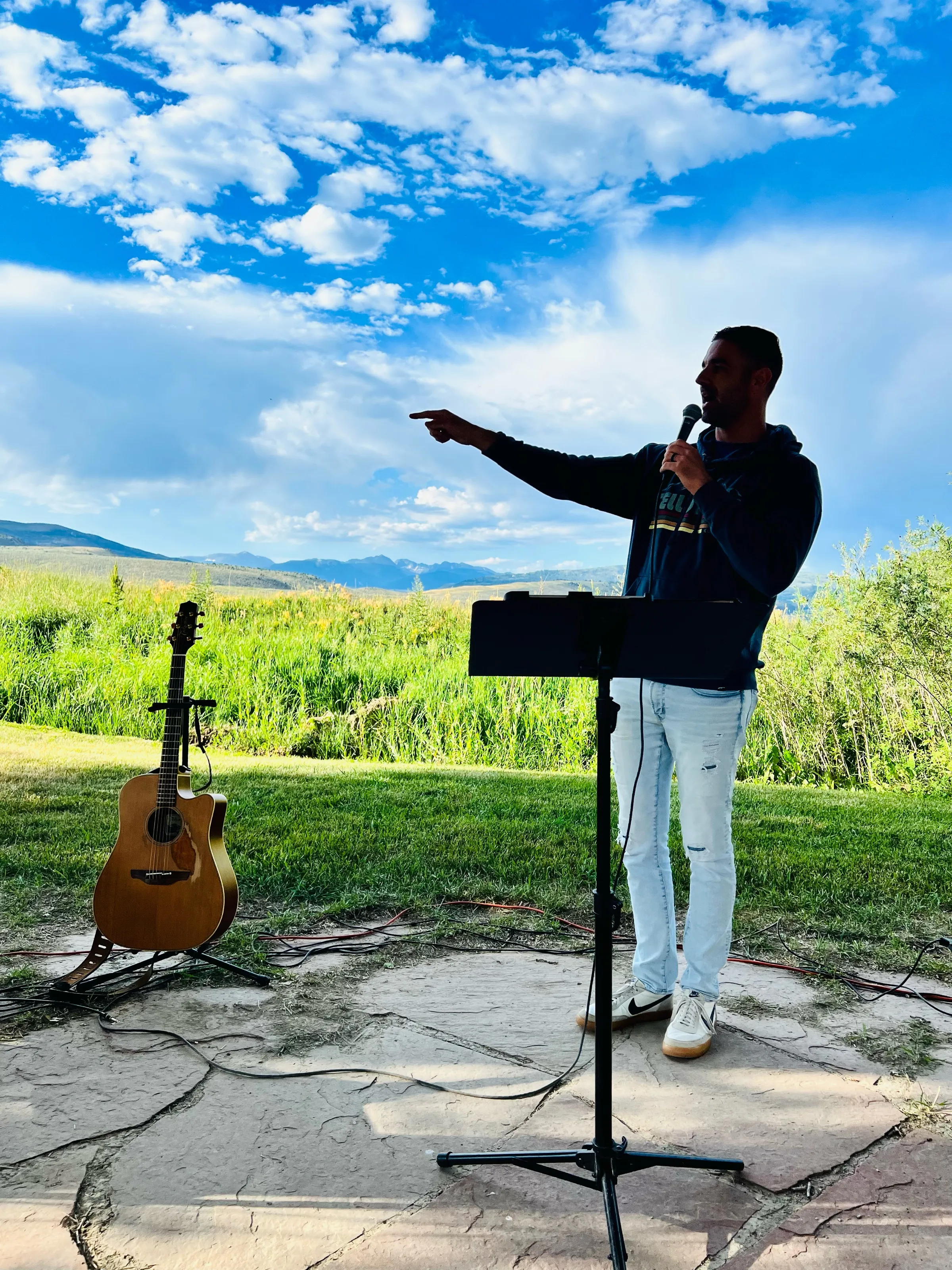 A person speaking into a microphone points outward beside an acoustic guitar and music stand, with mountains behind.