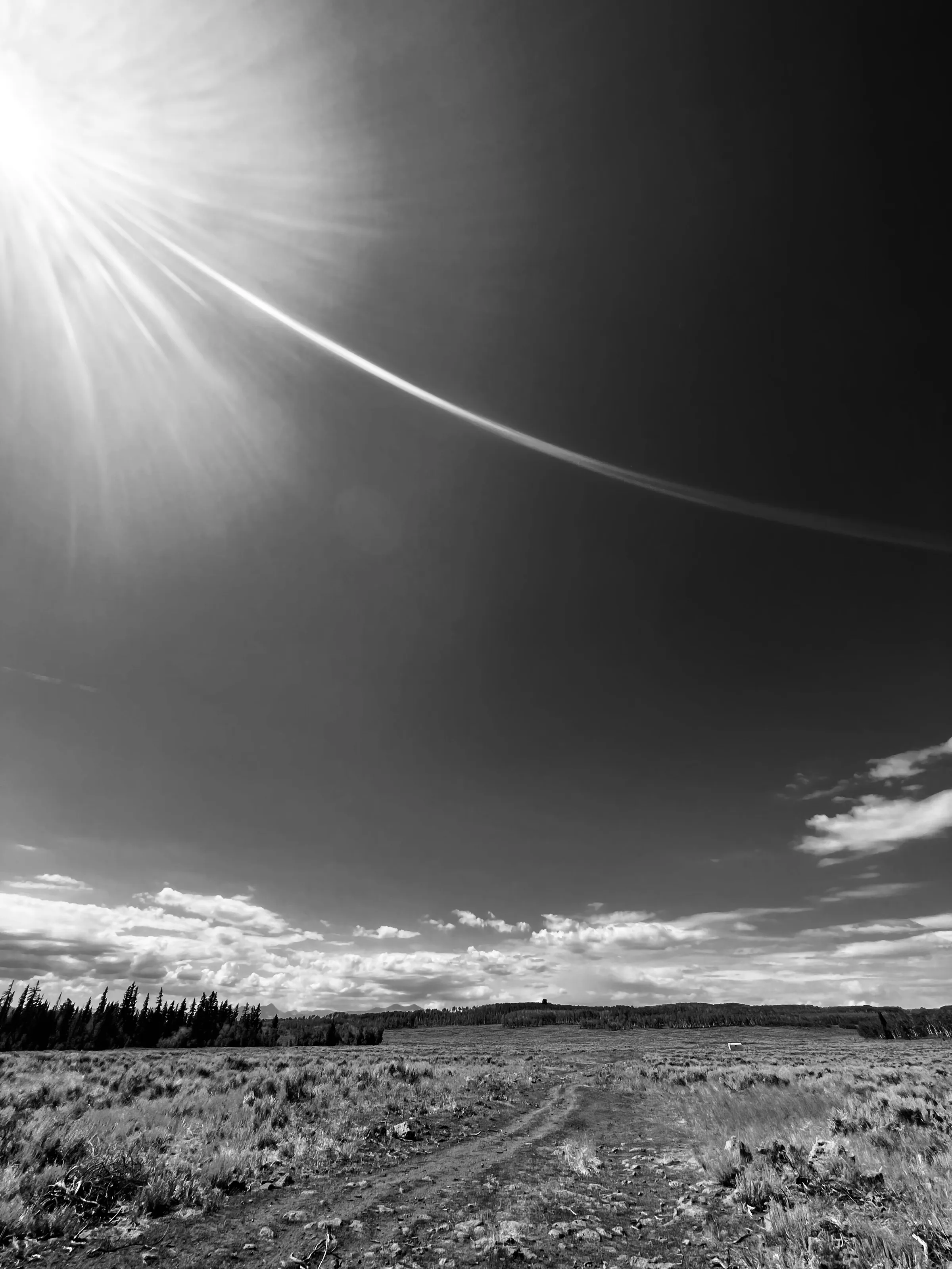 Black-and-white landscape with a bright sun at upper left, a diagonal lens flare, low clouds, and a rough field below.