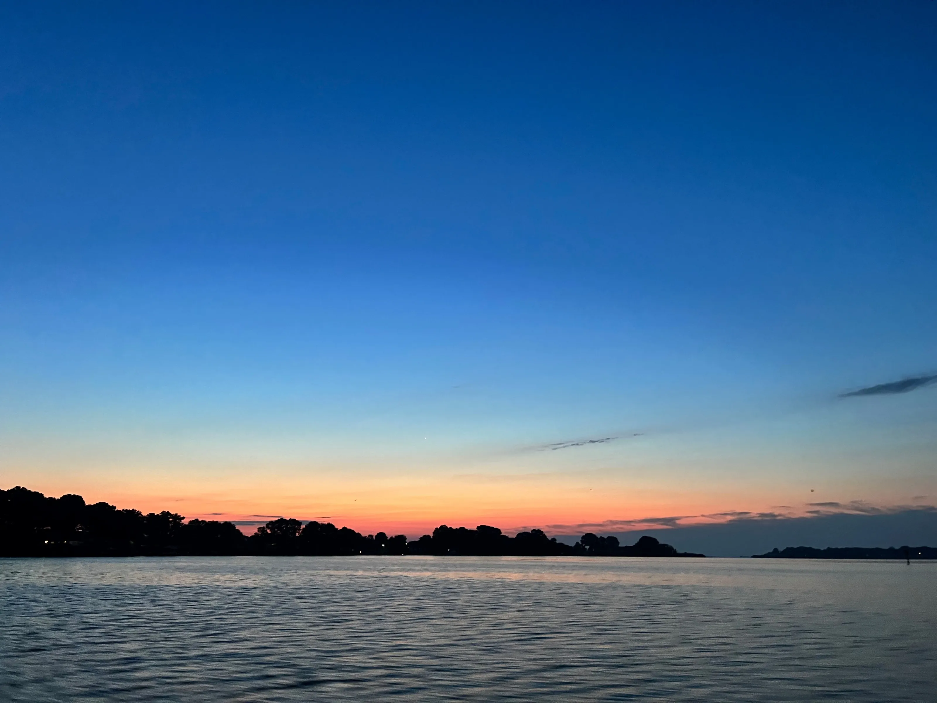A broad blue evening sky over calm water and a dark low shoreline, with a narrow orange band at the horizon.