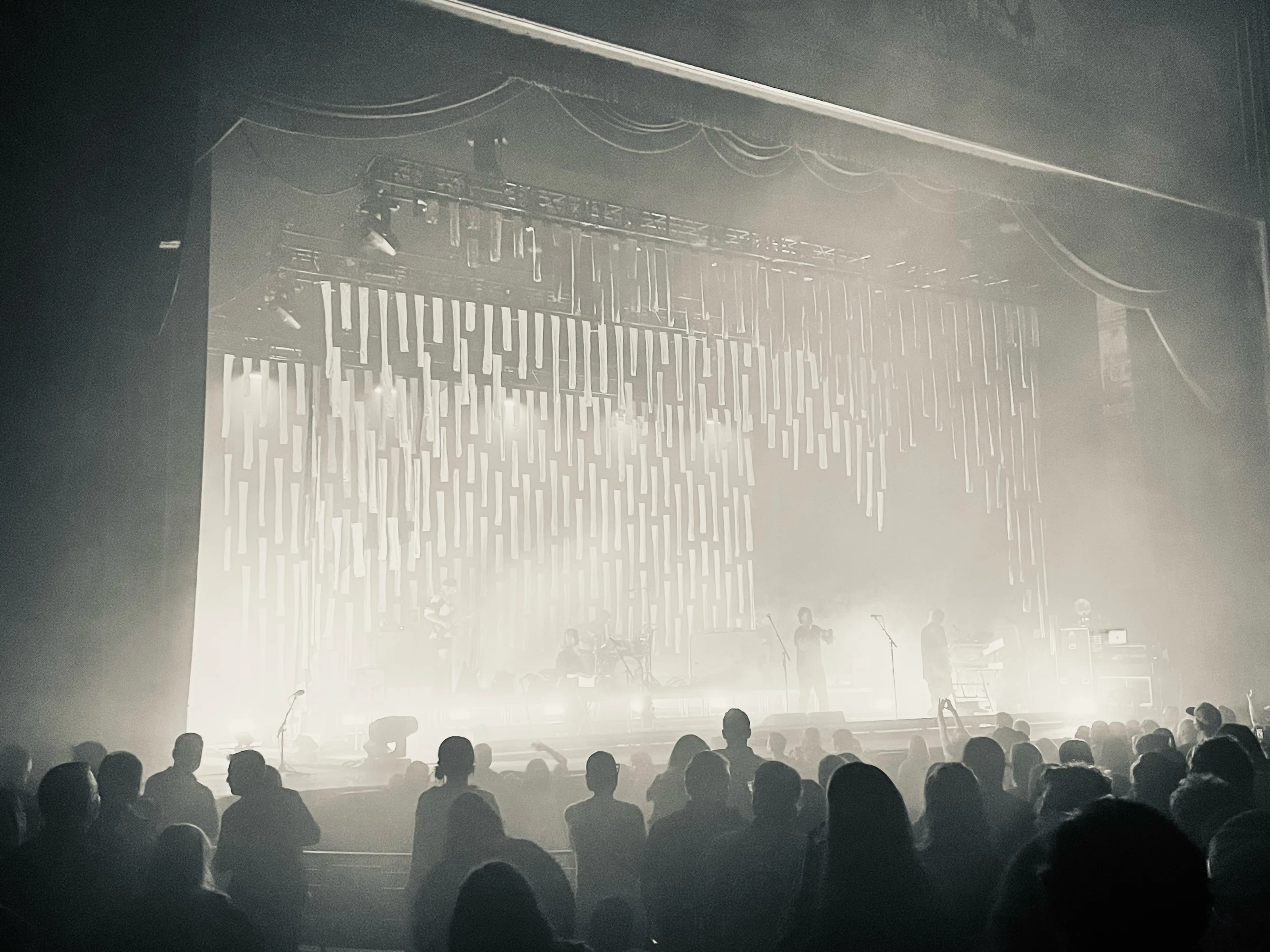 Silhouetted concertgoers face a bright fog-filled stage with hanging vertical lights.