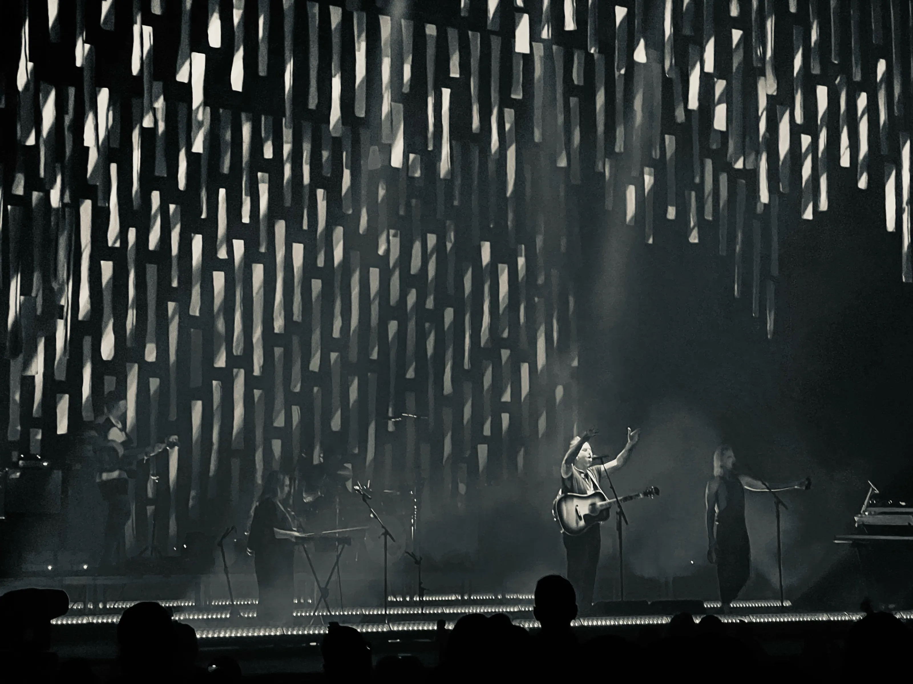 Dark concert stage with several performers silhouetted beneath hundreds of hanging rectangular light elements.
