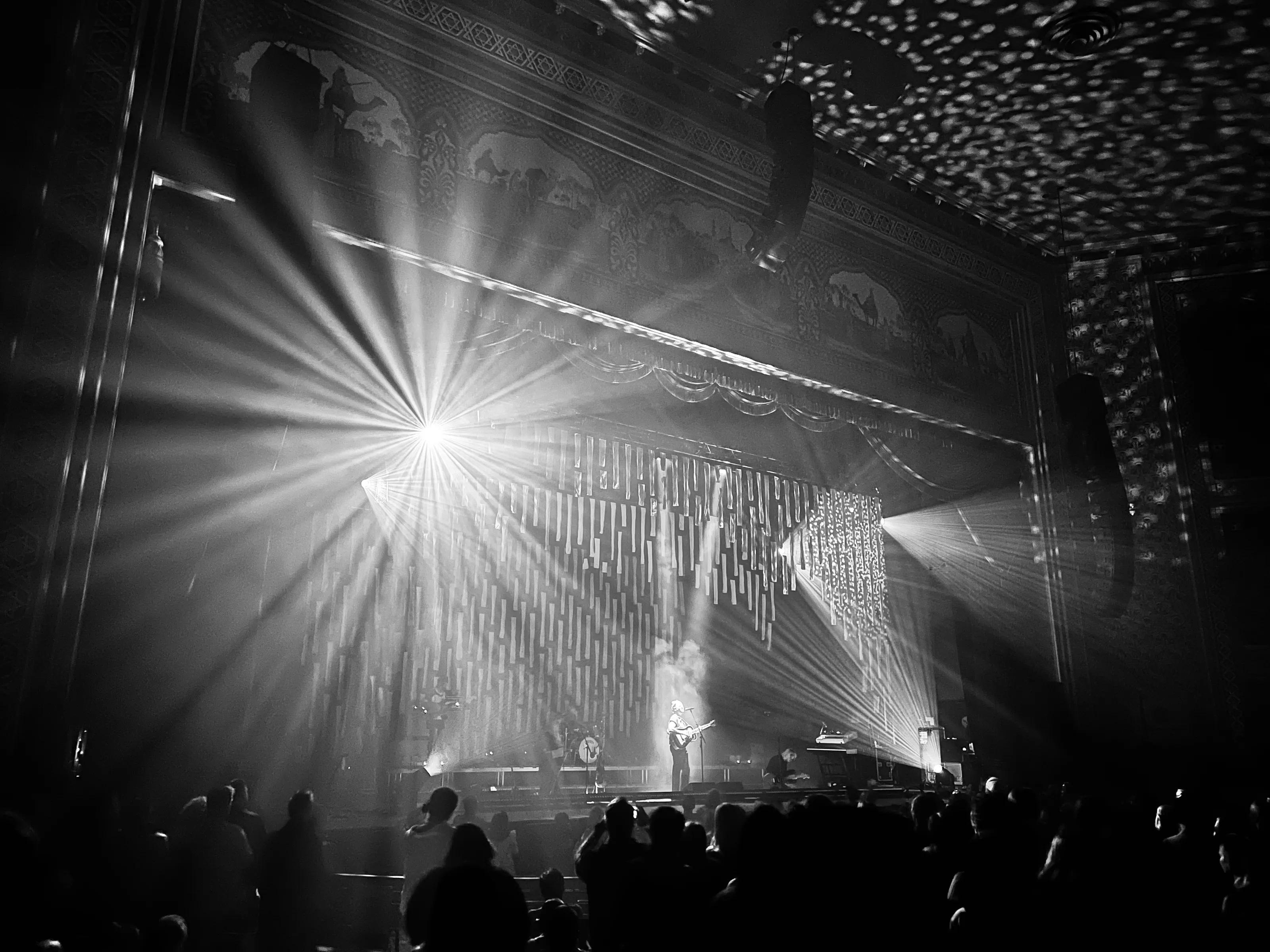 Black-and-white concert scene with bright rays of stage light, a small performer, and a crowd in silhouette.