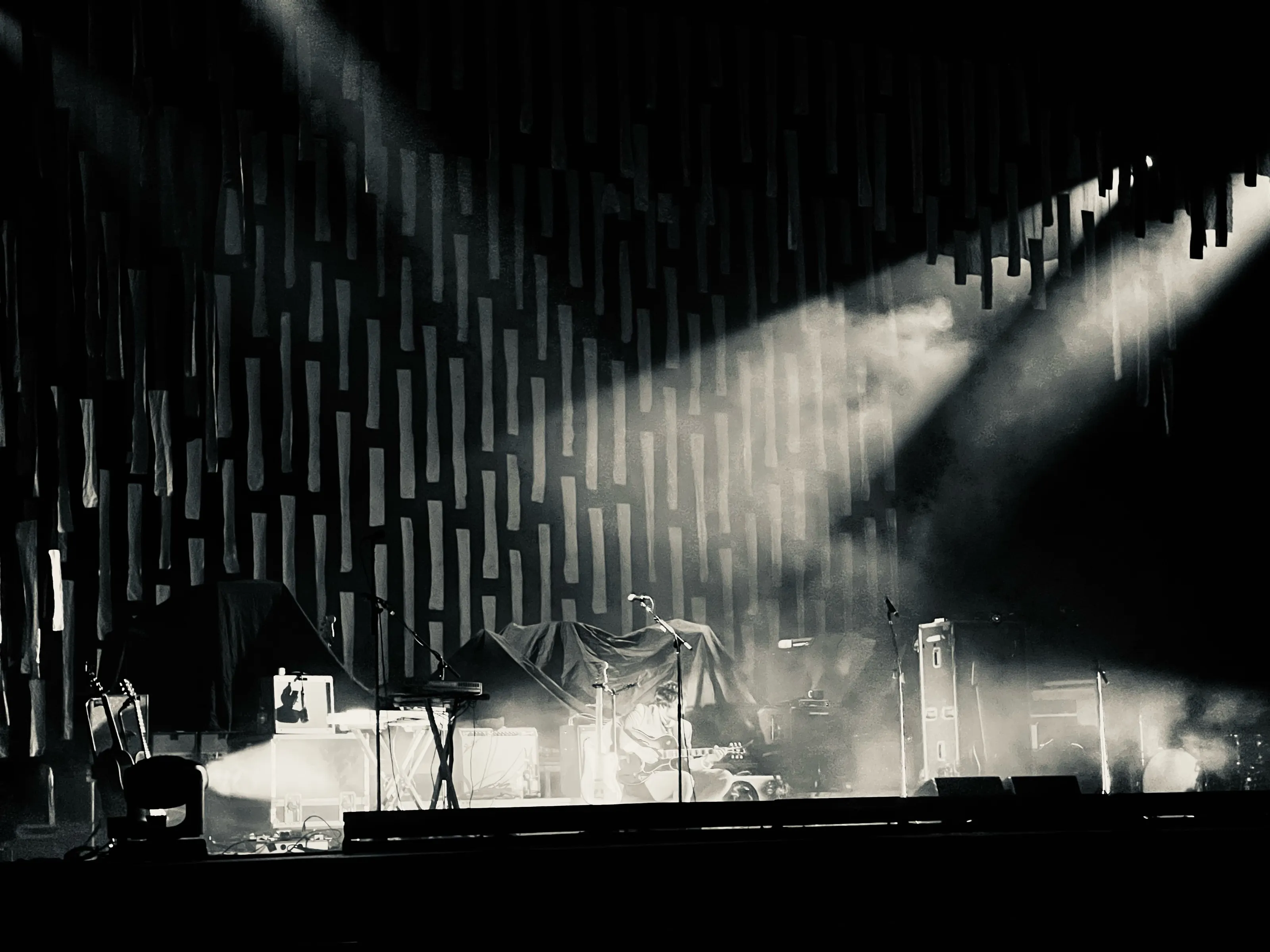 Bright beams of stage light shine through haze onto covered instruments and equipment on a dark stage.