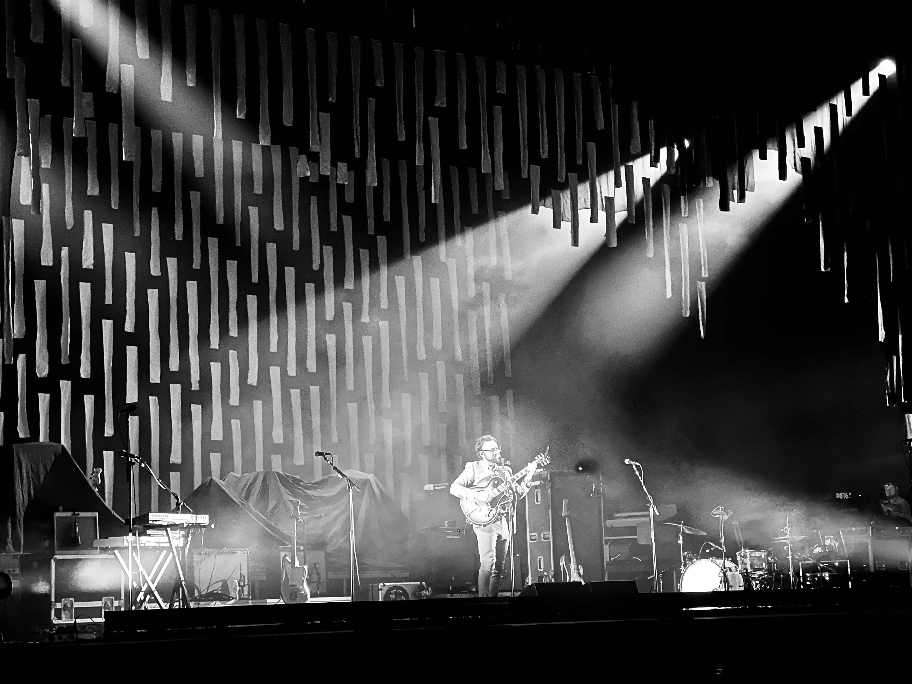 Black-and-white concert photograph of a guitarist on a large stage beneath rows of hanging rectangular elements and bright diagonal beams of light.