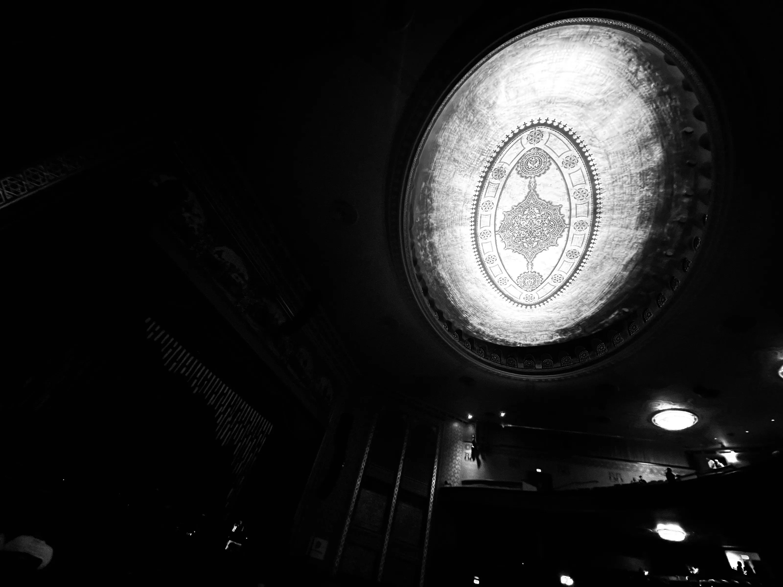 Black-and-white view upward in a dark theater, with a bright oval ornamental ceiling medallion and small lights around it.