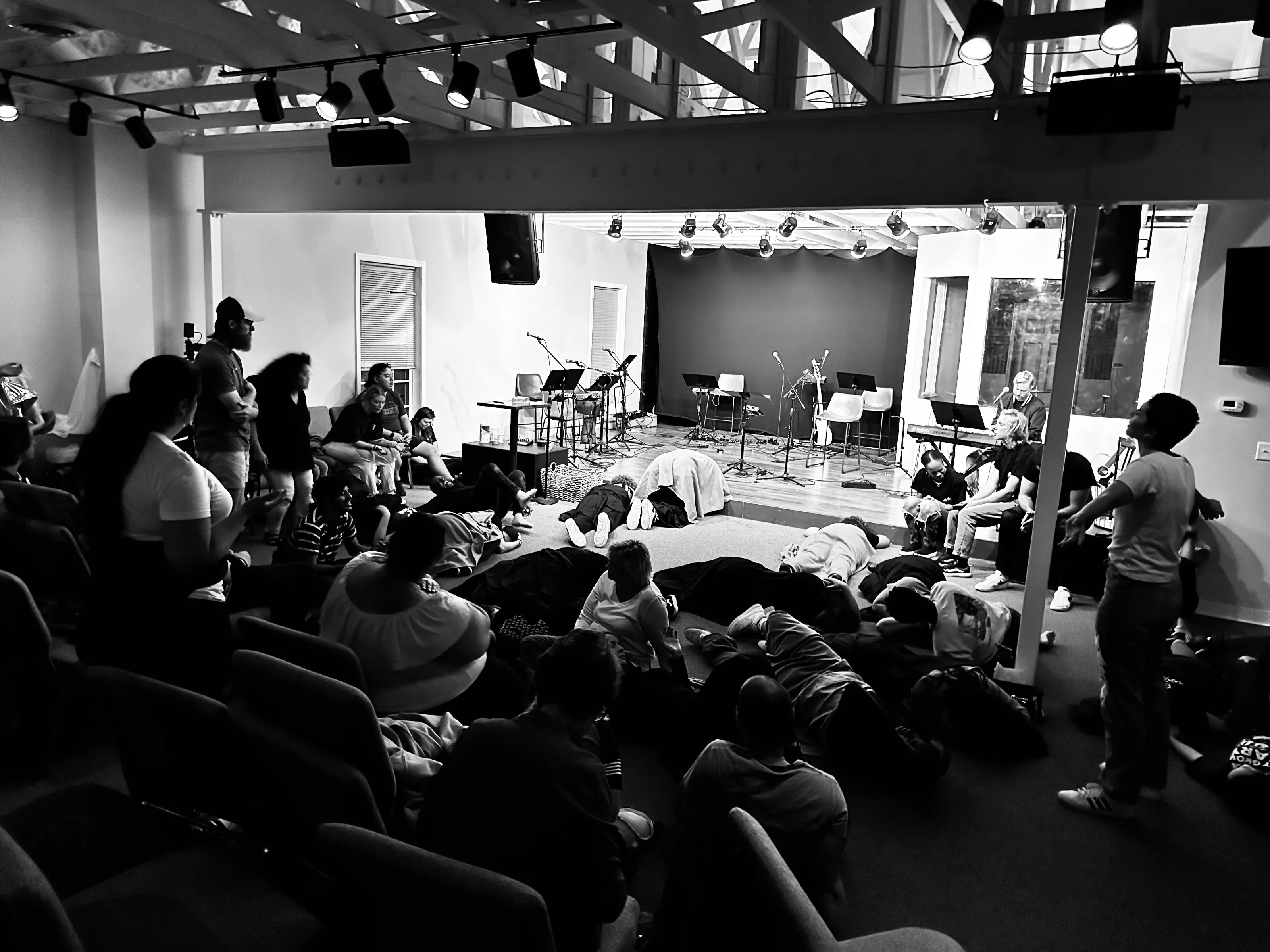 A black-and-white wide view of people lying or sitting in a small room facing a performance area.