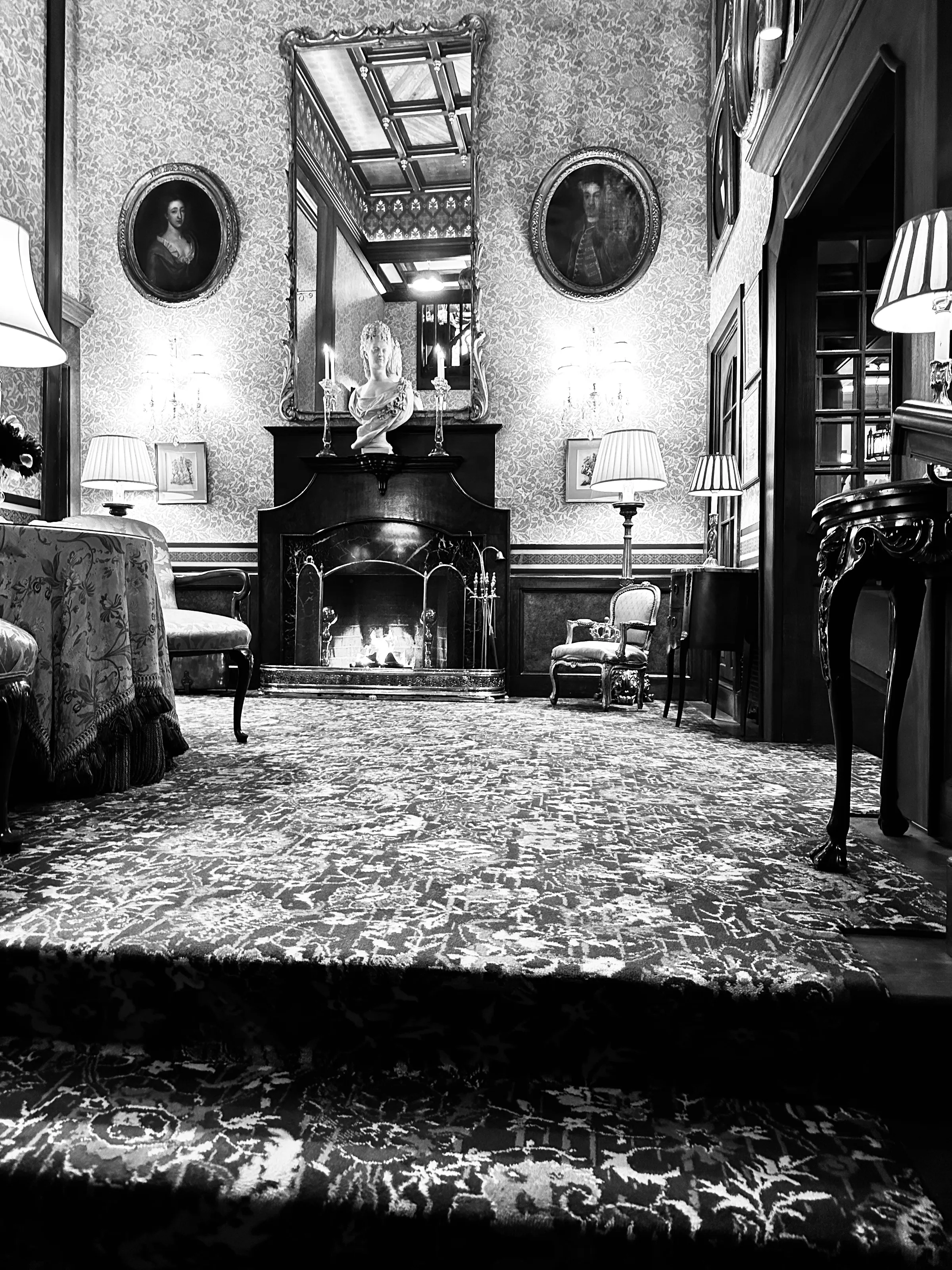 A black-and-white view from a carpeted stair into an ornate room with a fireplace, mirror, and lamps.