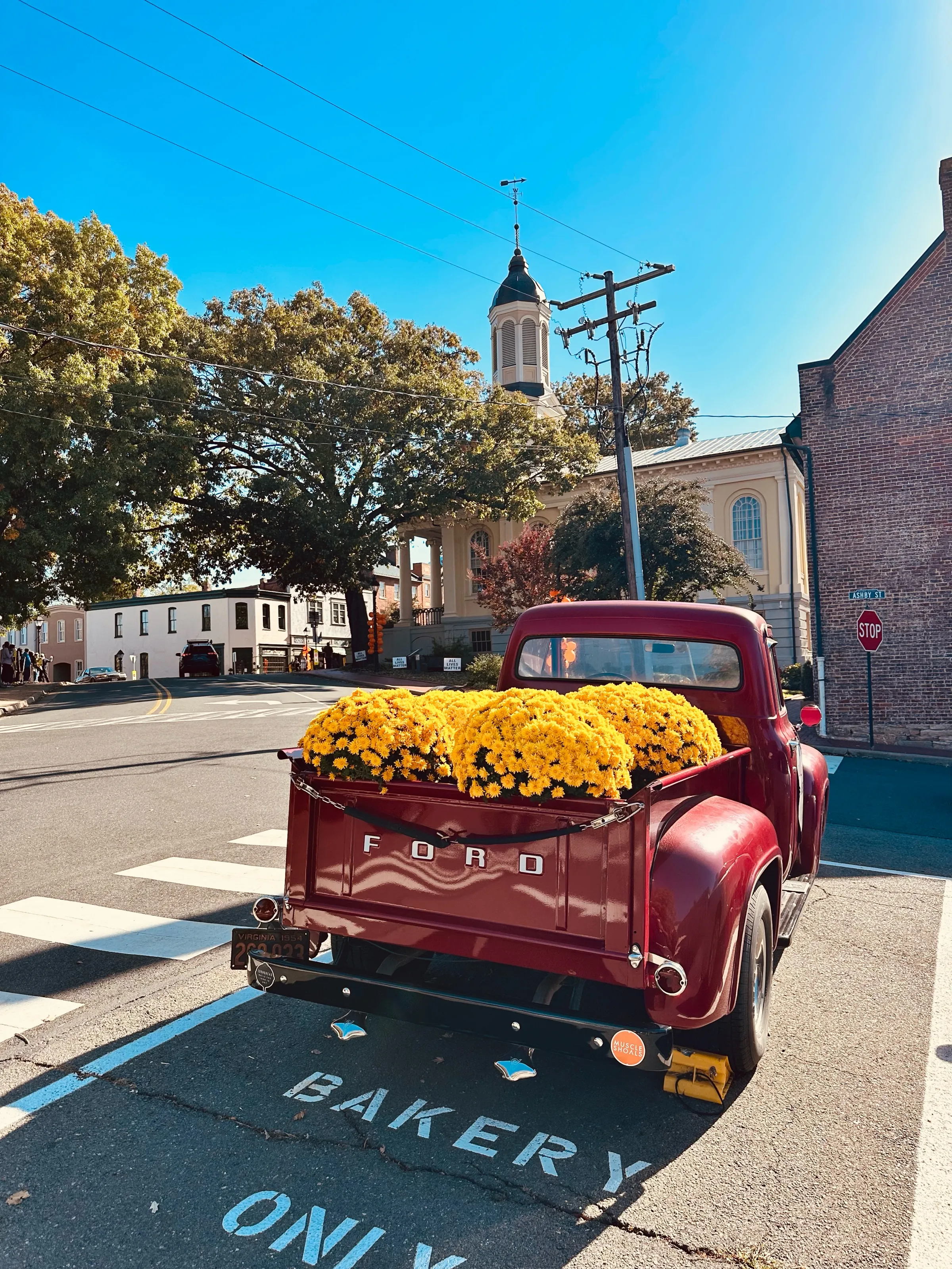 A red vintage Ford pickup truck filled with yellow flowers is parked near a church and a street intersection.