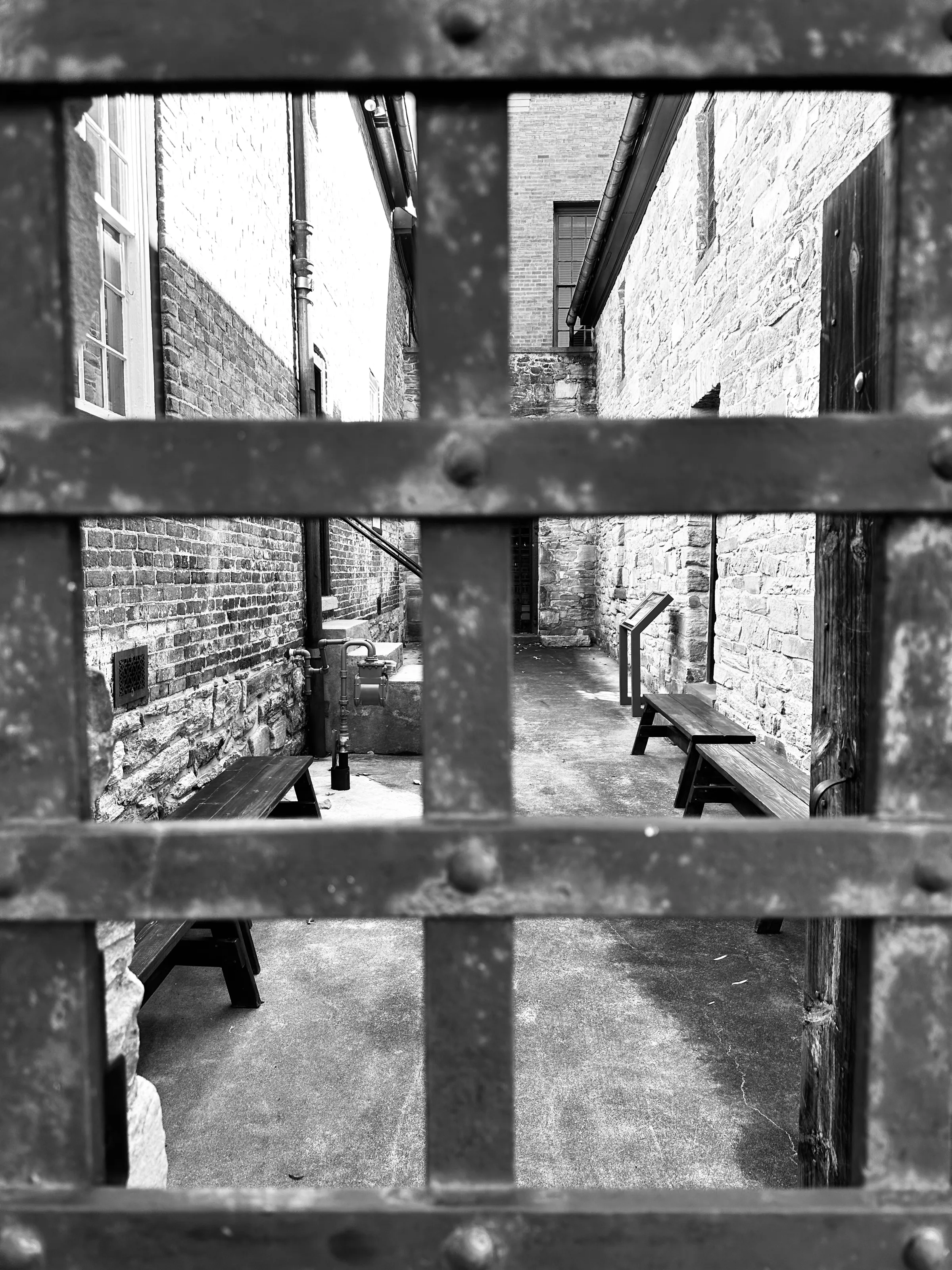 Black-and-white photograph of benches in a narrow stone courtyard viewed through a metal gate grid.