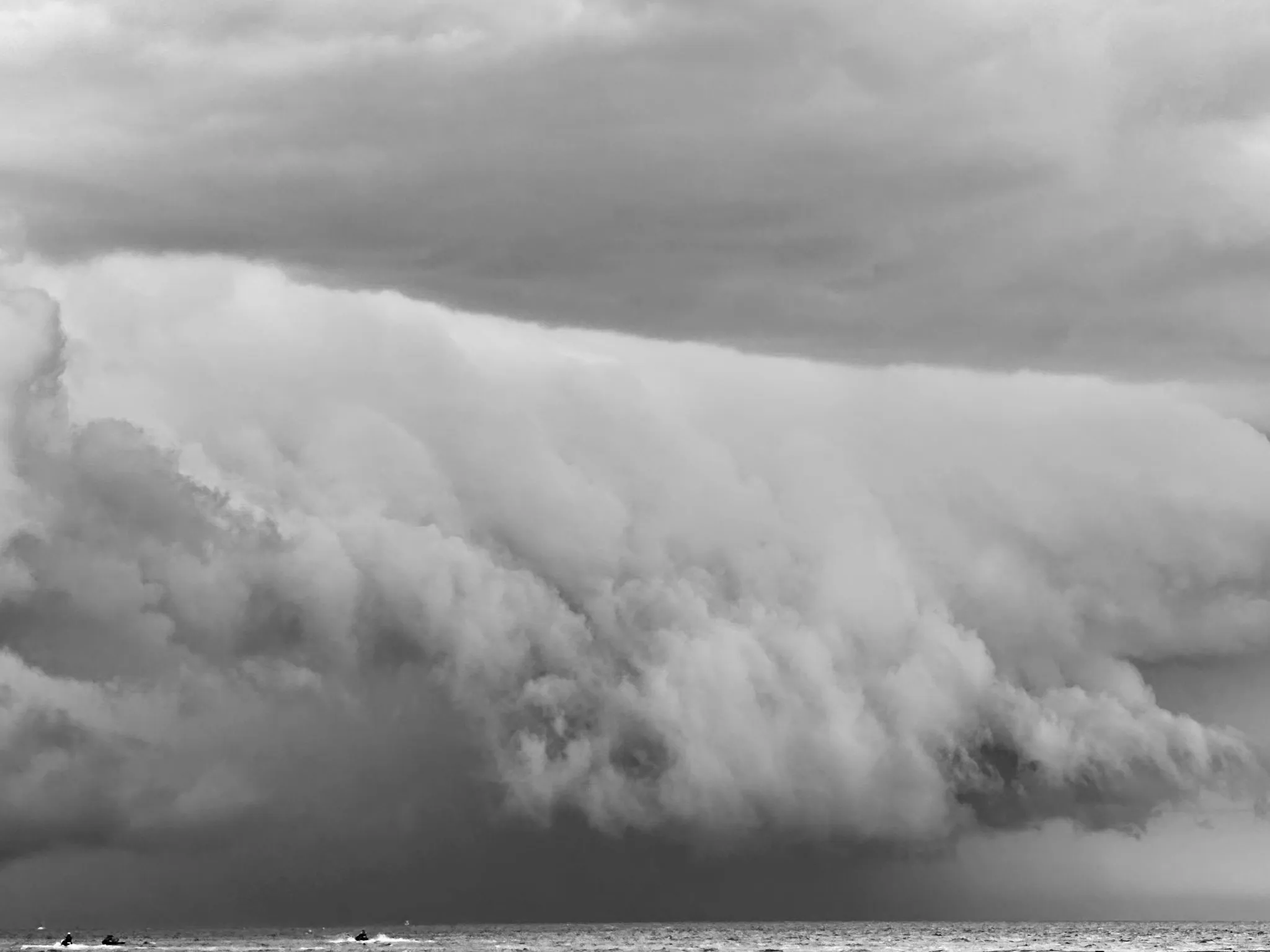 Black-and-white photograph of a thick, rolling mass of storm clouds above a narrow strip of ocean.