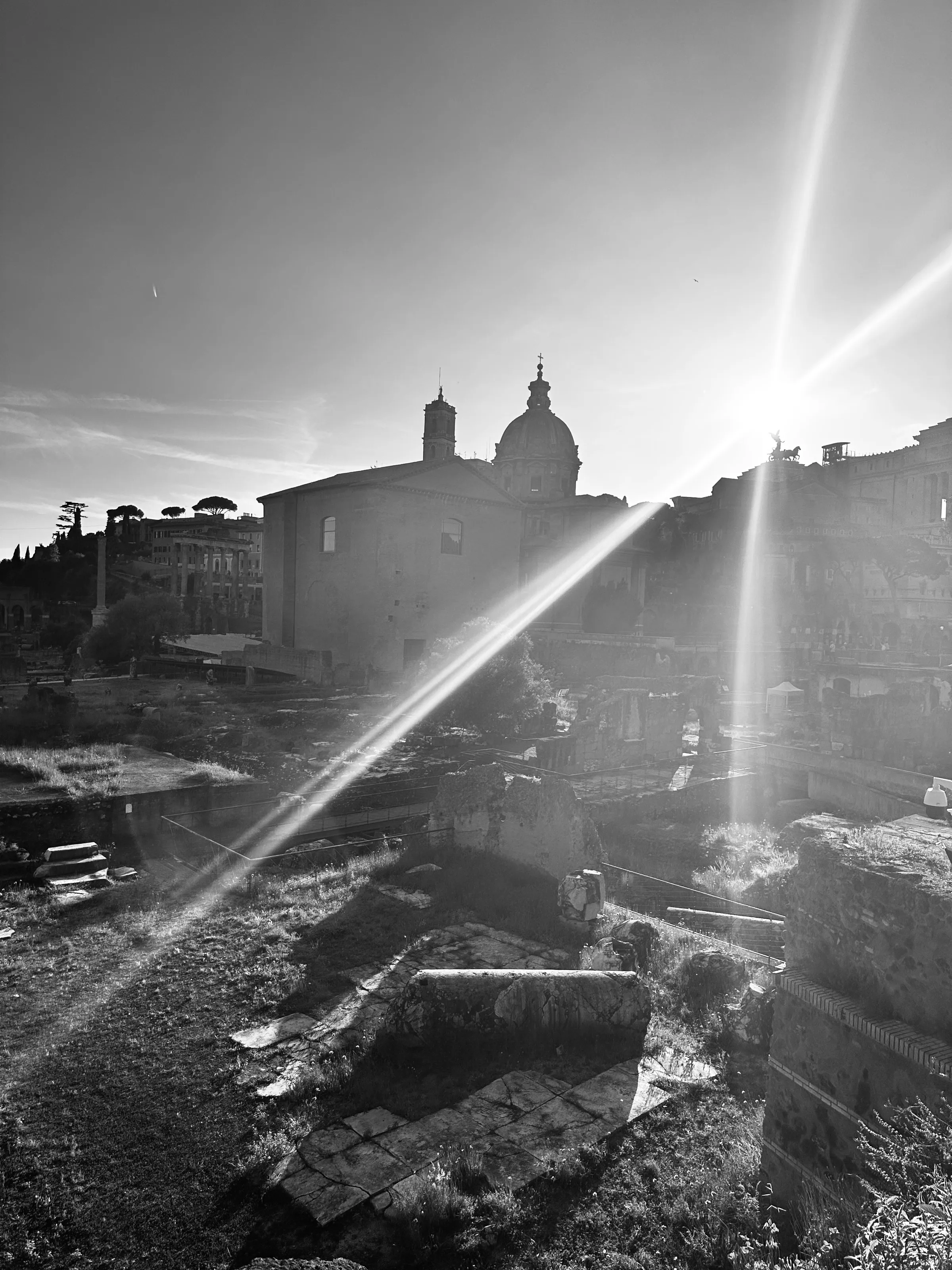 Black-and-white view of ancient stone ruins and domed church buildings with bright sun flare crossing the frame.