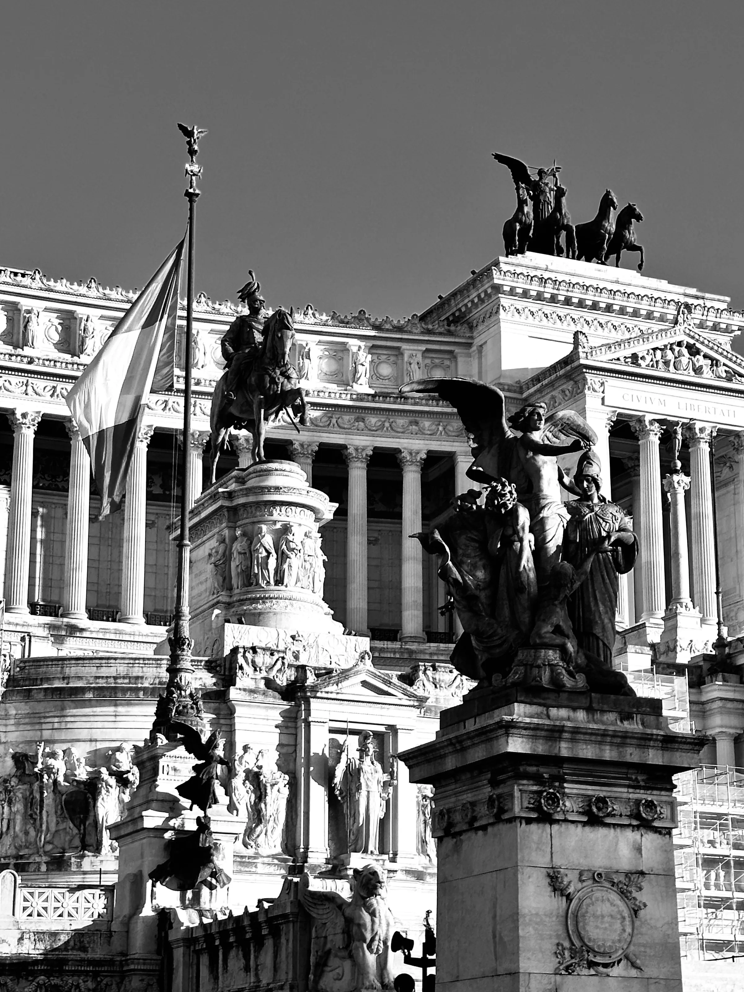 Large statues and columns stand in front of an ornate monumental building with an Italian flag.
