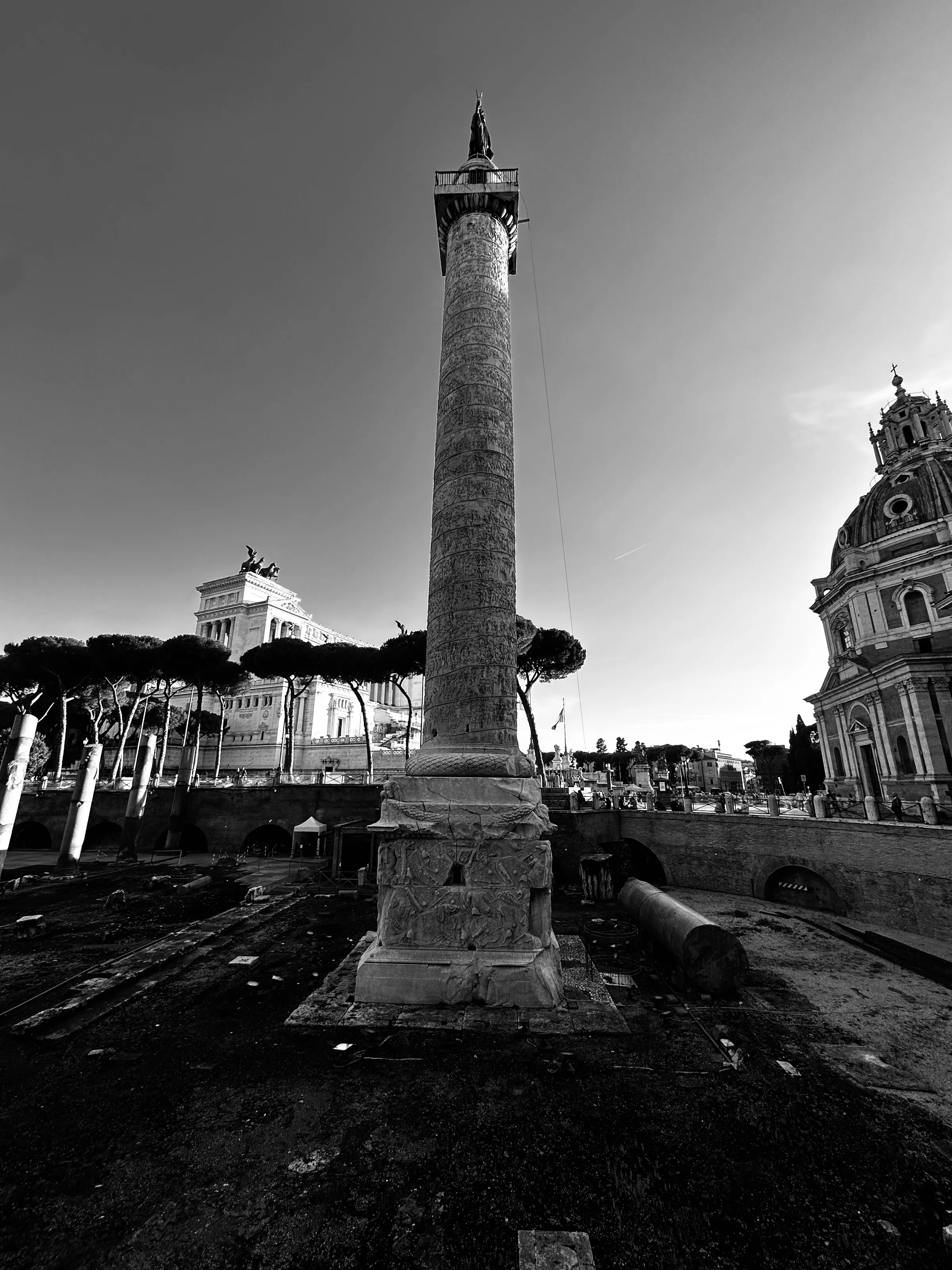 Black-and-white low-angle view of a tall stone column in ruins, with a church dome on the right and umbrella pines on the left.