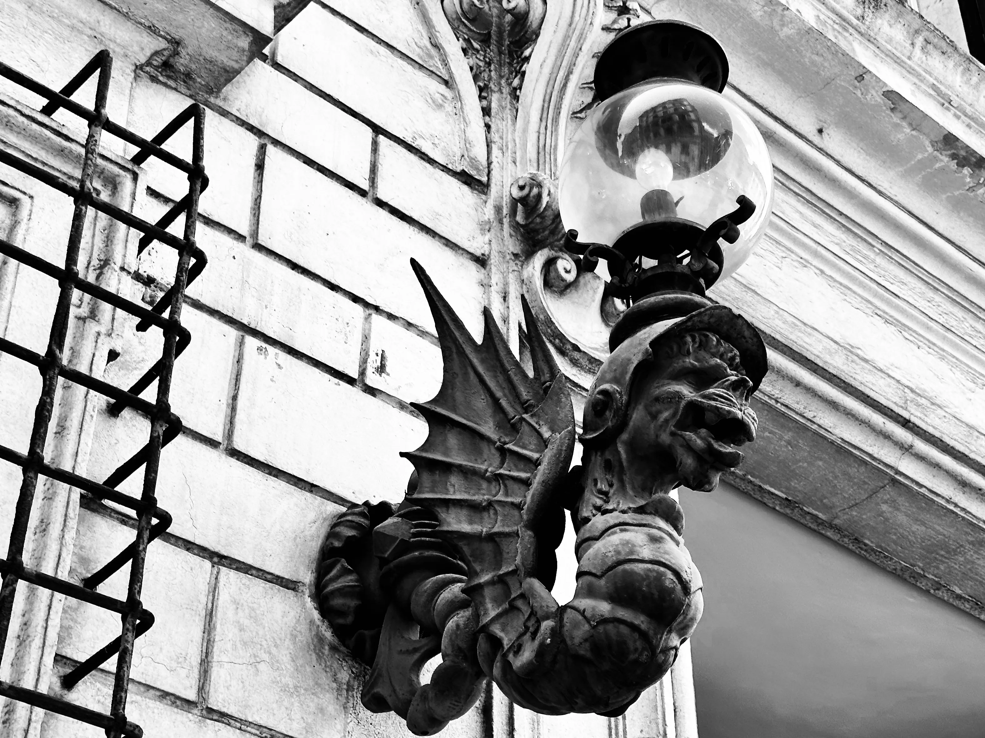 Black-and-white close view of a winged gargoyle-like sculpture holding a round glass lamp on a weathered building facade.