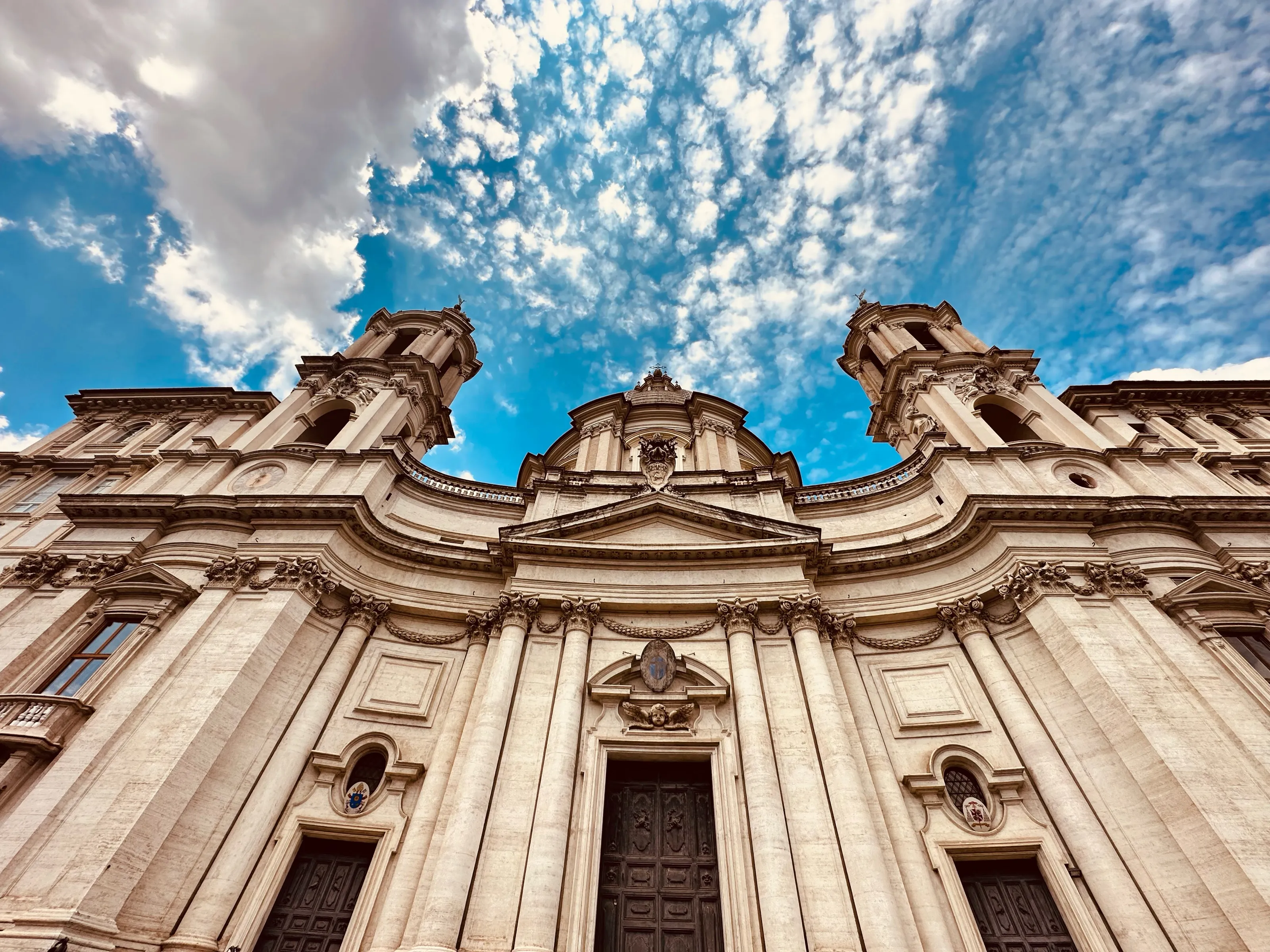 A low-angle view of an elaborate pale stone church facade with two towers against a bright blue sky and textured clouds.