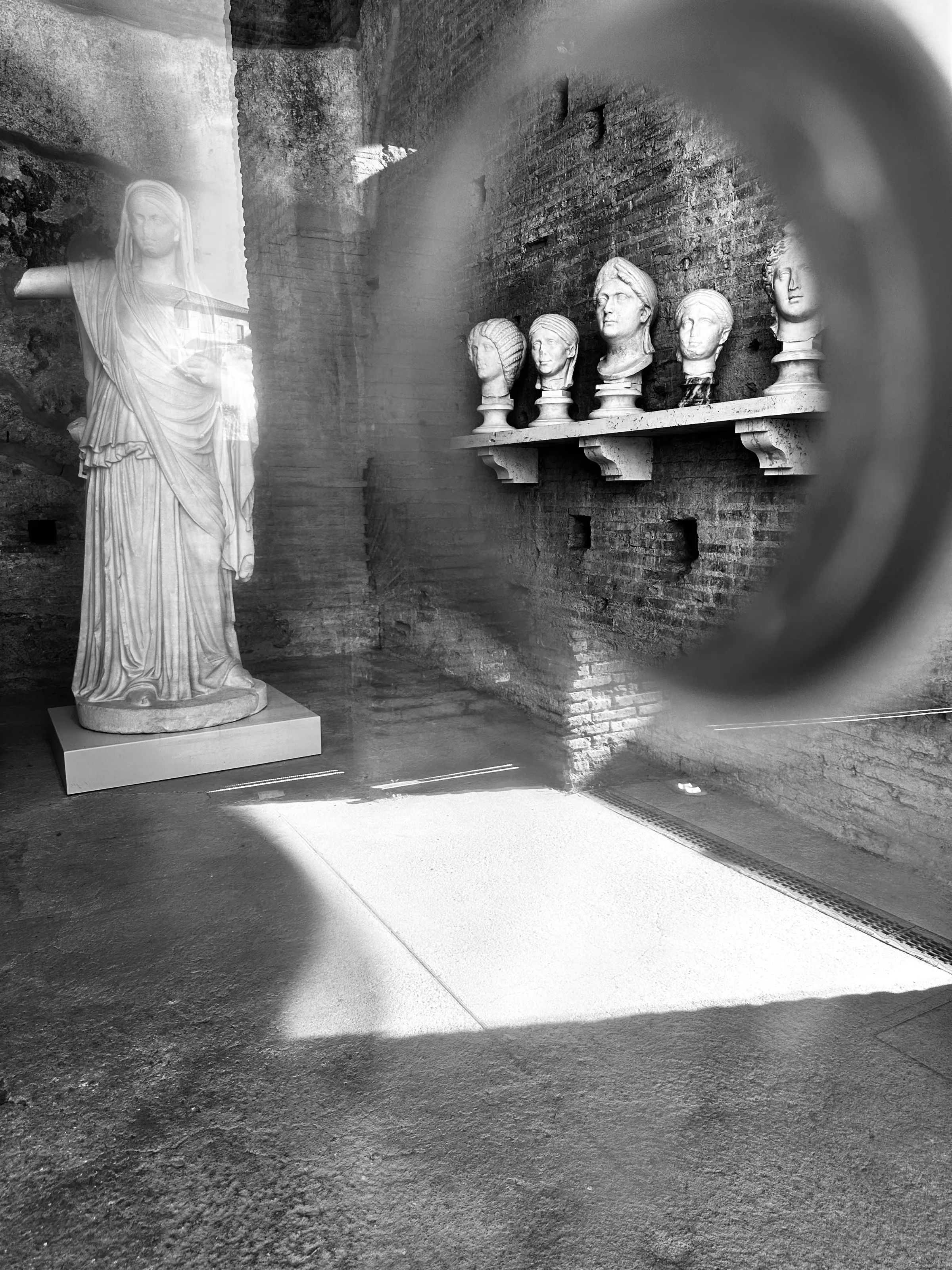 Black-and-white view of a robed statue beside a shelf of sculpted heads, seen through a circular reflection.