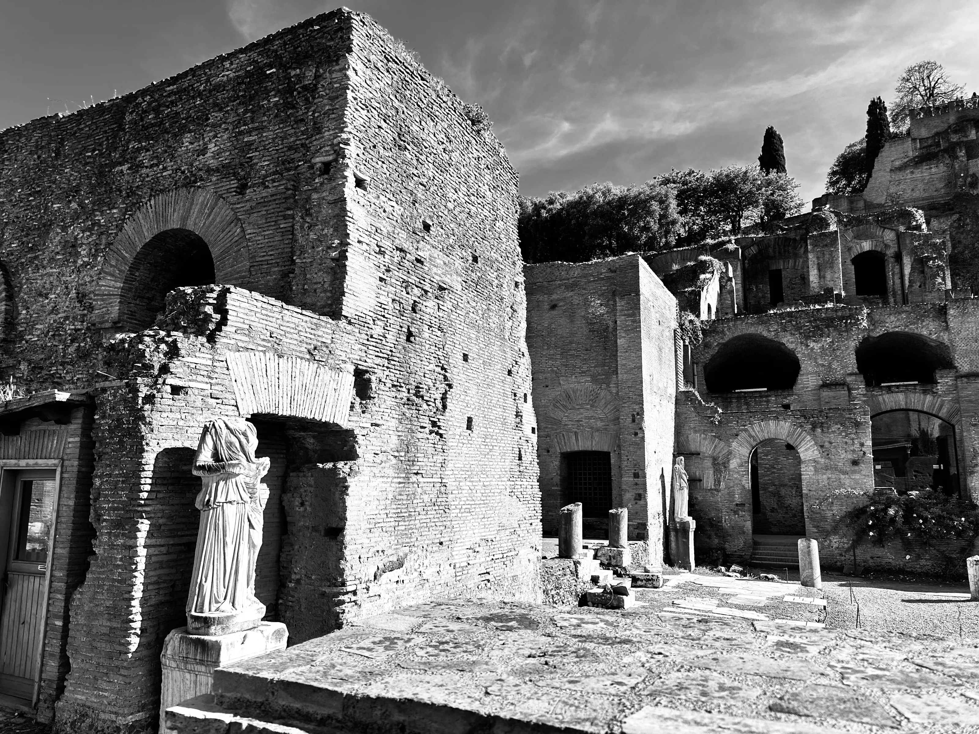 A draped statue stands beside weathered brick ruins and arched openings in a black-and-white photograph.