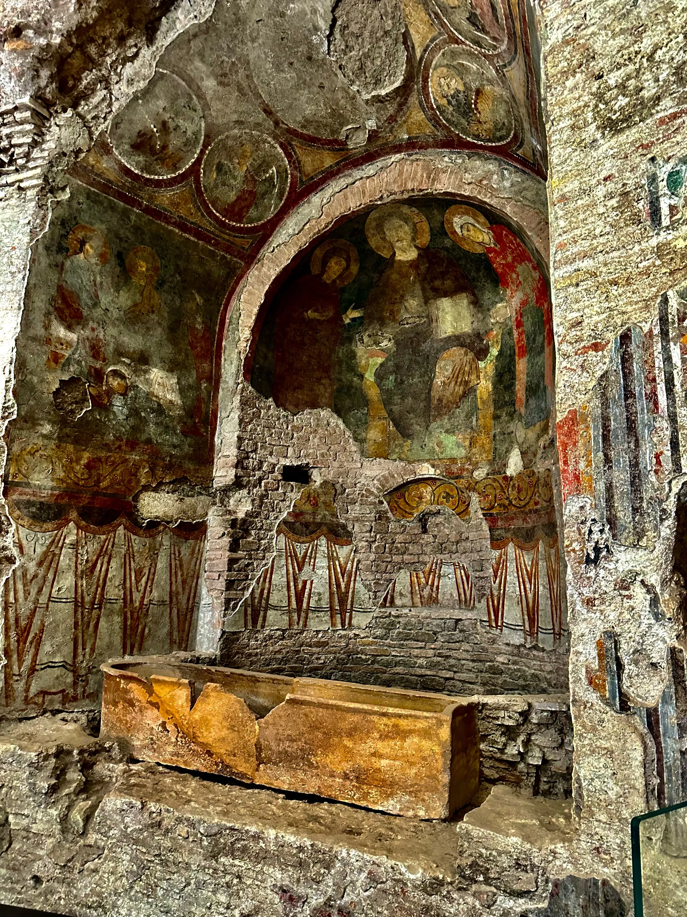 A weathered stone interior with faded religious frescoes, an arched opening, and a large stone coffin.