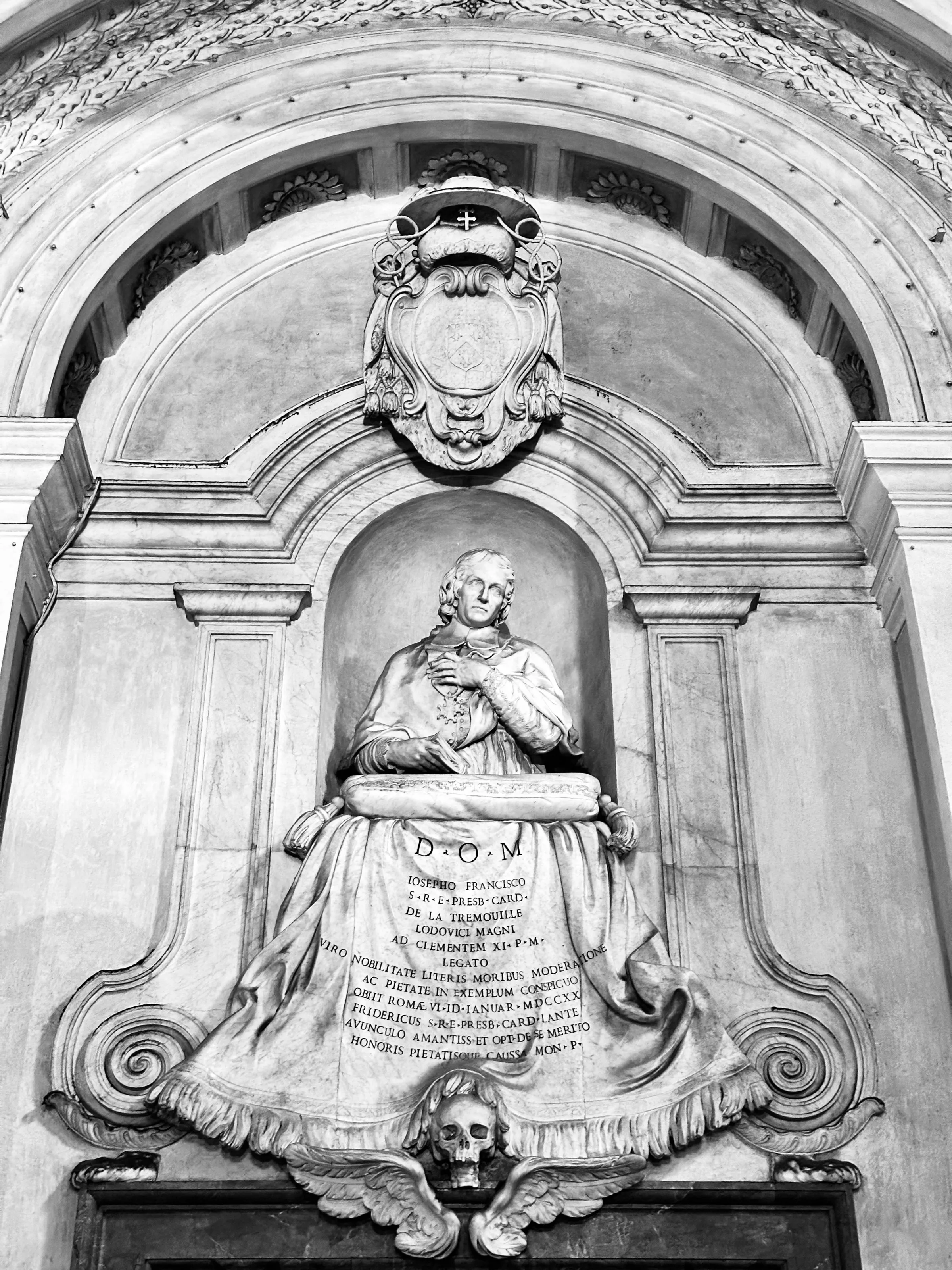 Black-and-white photograph of a carved marble memorial showing a seated figure above an inscription, draped stonework, and a skull at the base.