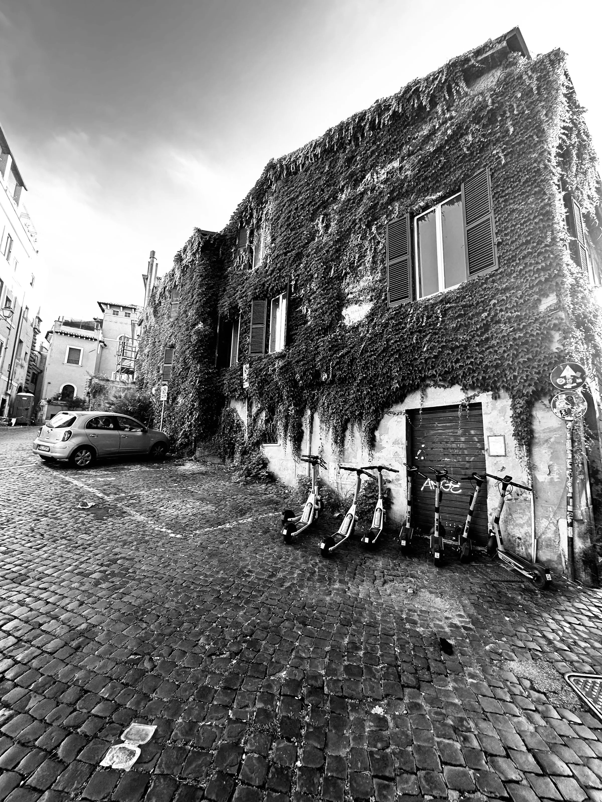 Three scooters lean beside a doorway in an ivy-covered building on a cobblestone street.