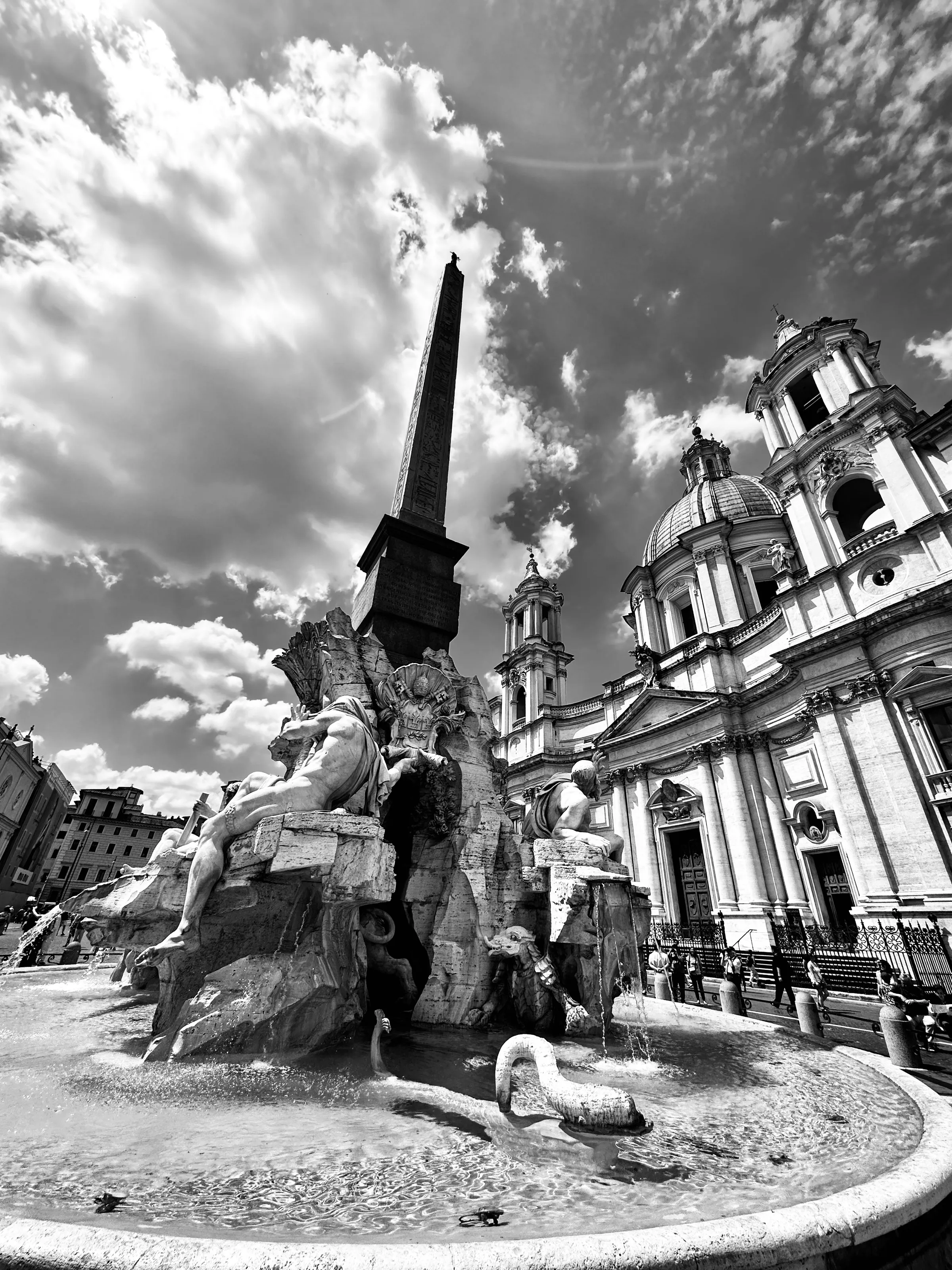 Black-and-white low-angle view of an obelisk, sculptural fountain, and ornate church facade, with small pedestrians in the background.