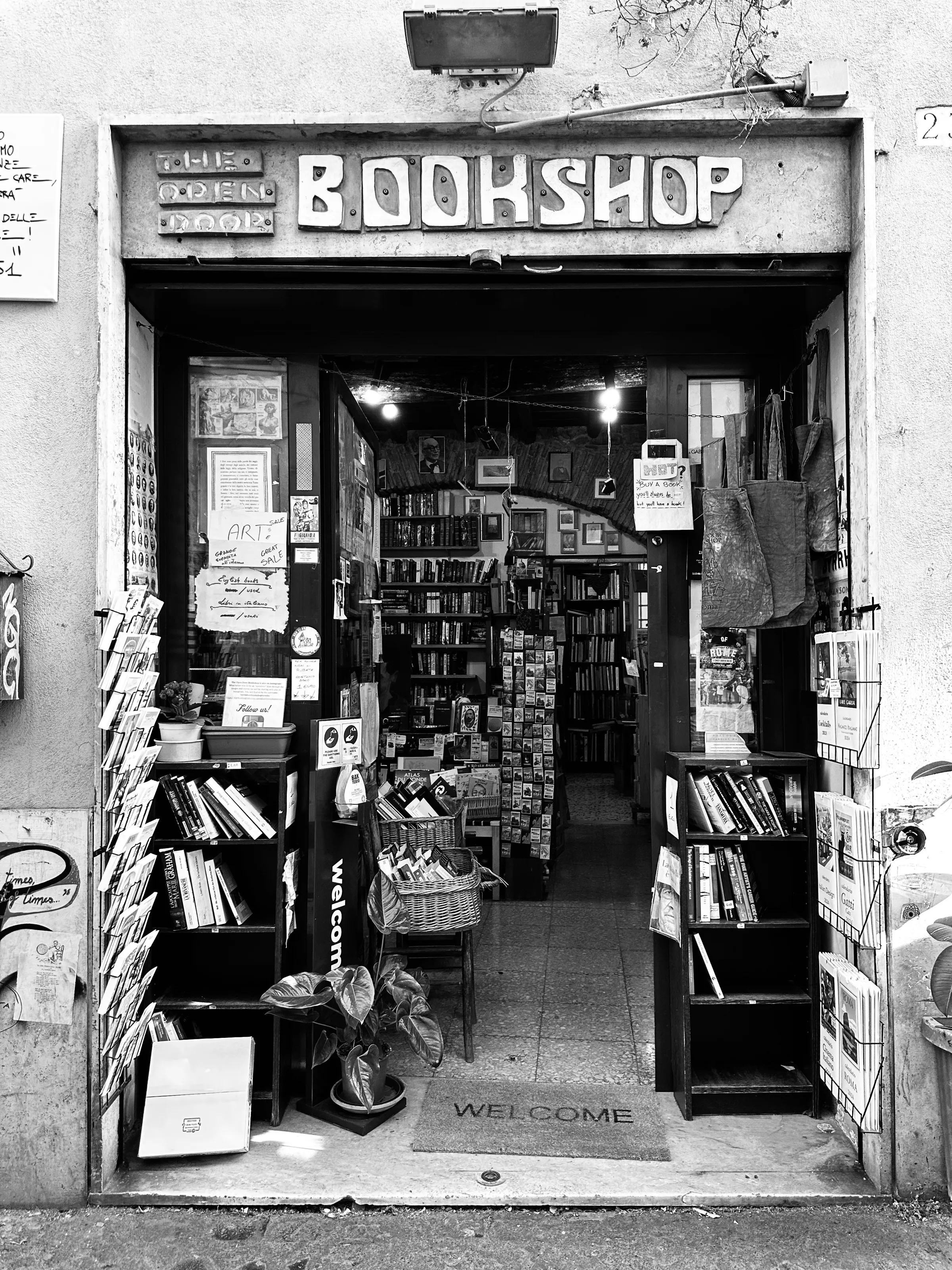 A black-and-white storefront of a small bookshop packed with shelves, books, signs, and postcards.