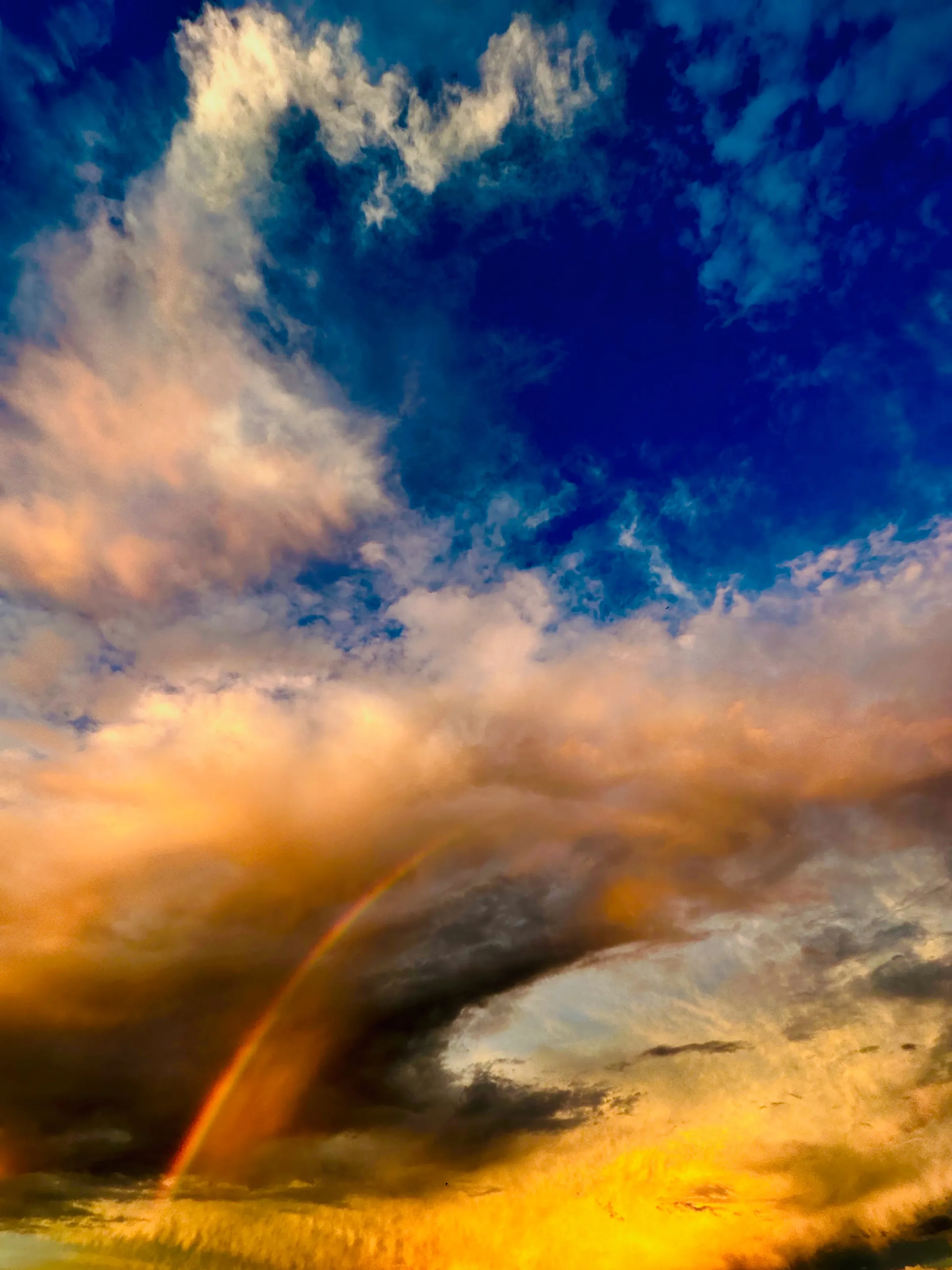 A partial rainbow rises from a dark storm cloud into orange and blue clouds over a bright horizon.