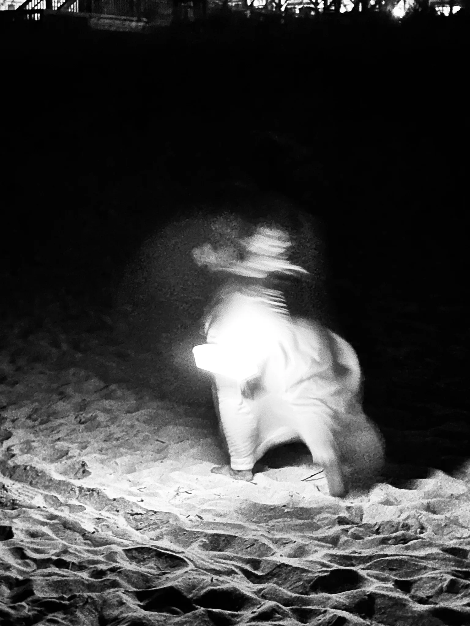 A blurred person standing on sand at night, lit by a bright flash against a nearly black background.
