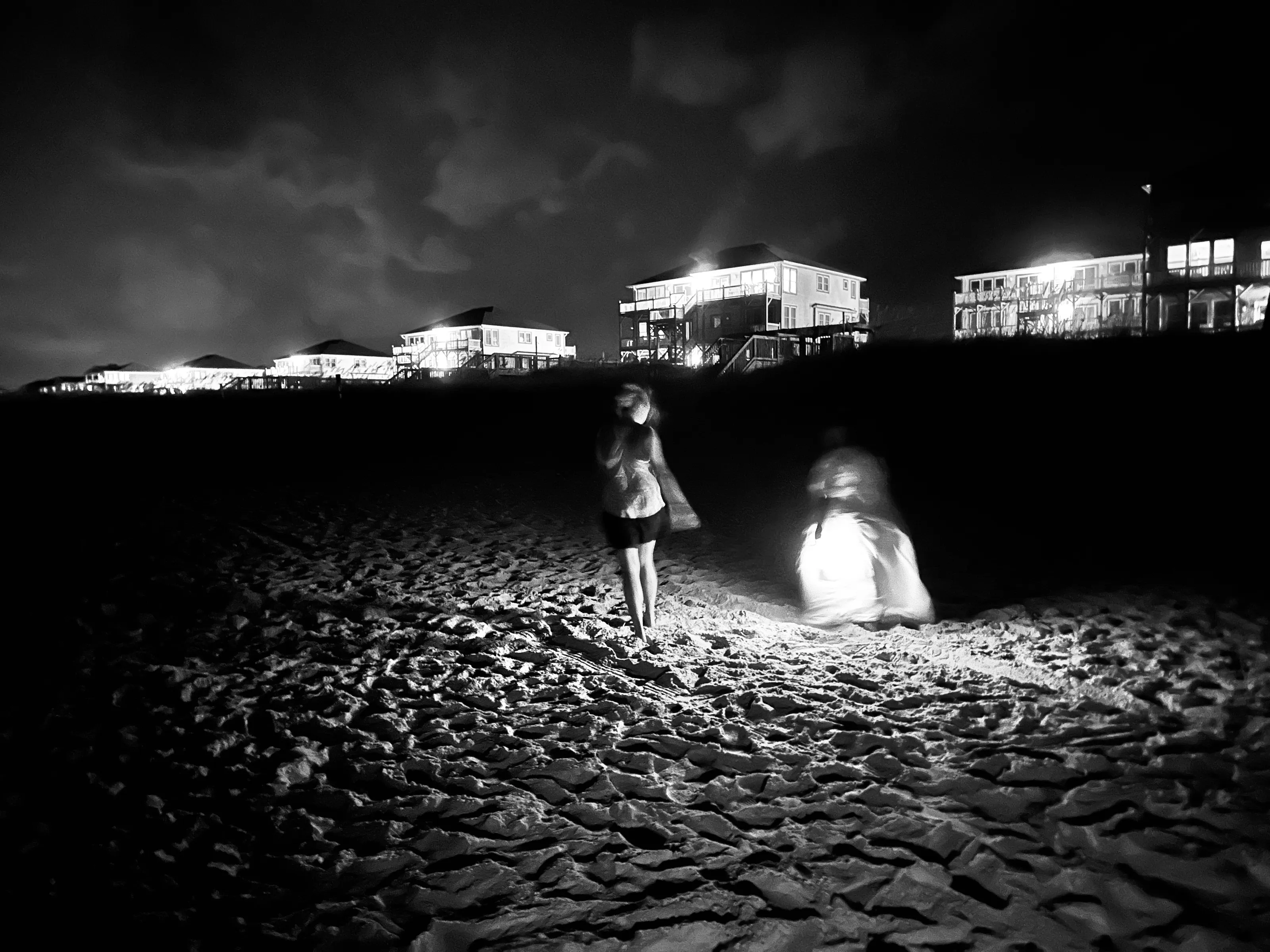 Black-and-white night photograph of two blurred people on a sandy beach beneath brightly lit houses.
