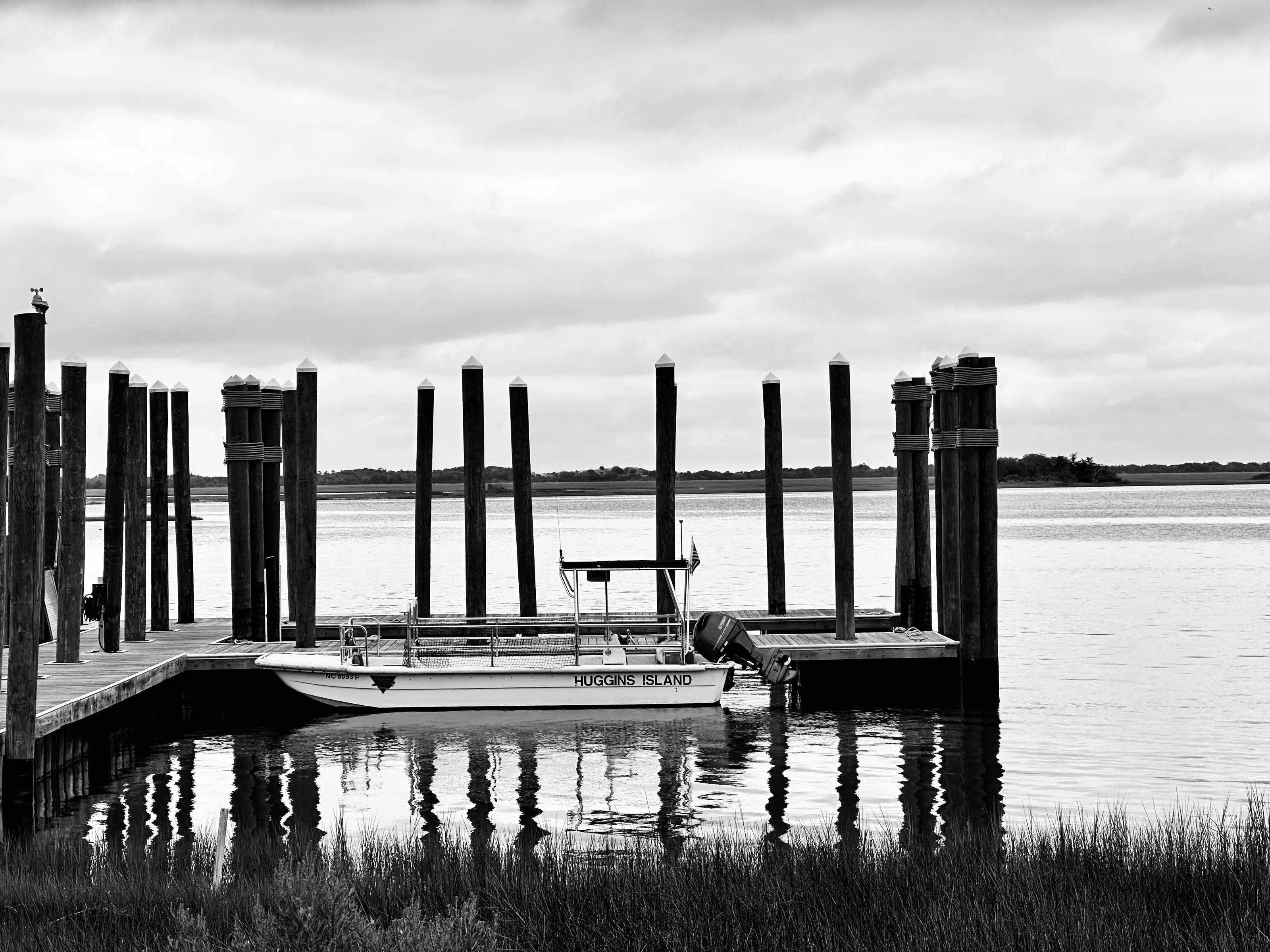 Black-and-white photograph of a small motorboat tied between tall dock pilings, with their reflections in calm water.