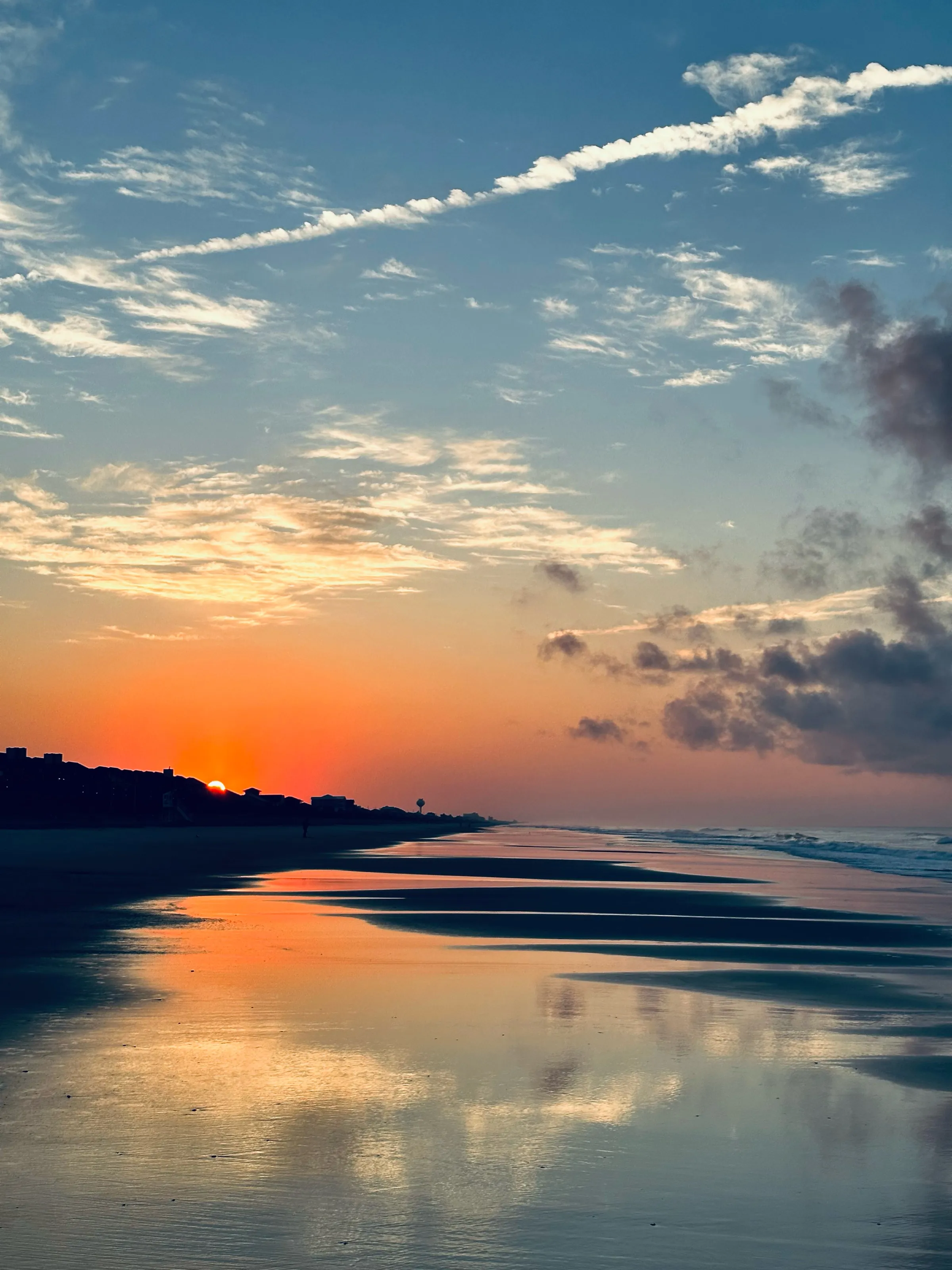 A sunset over the ocean reflected in wet sand, with dark shoreline silhouettes at left.