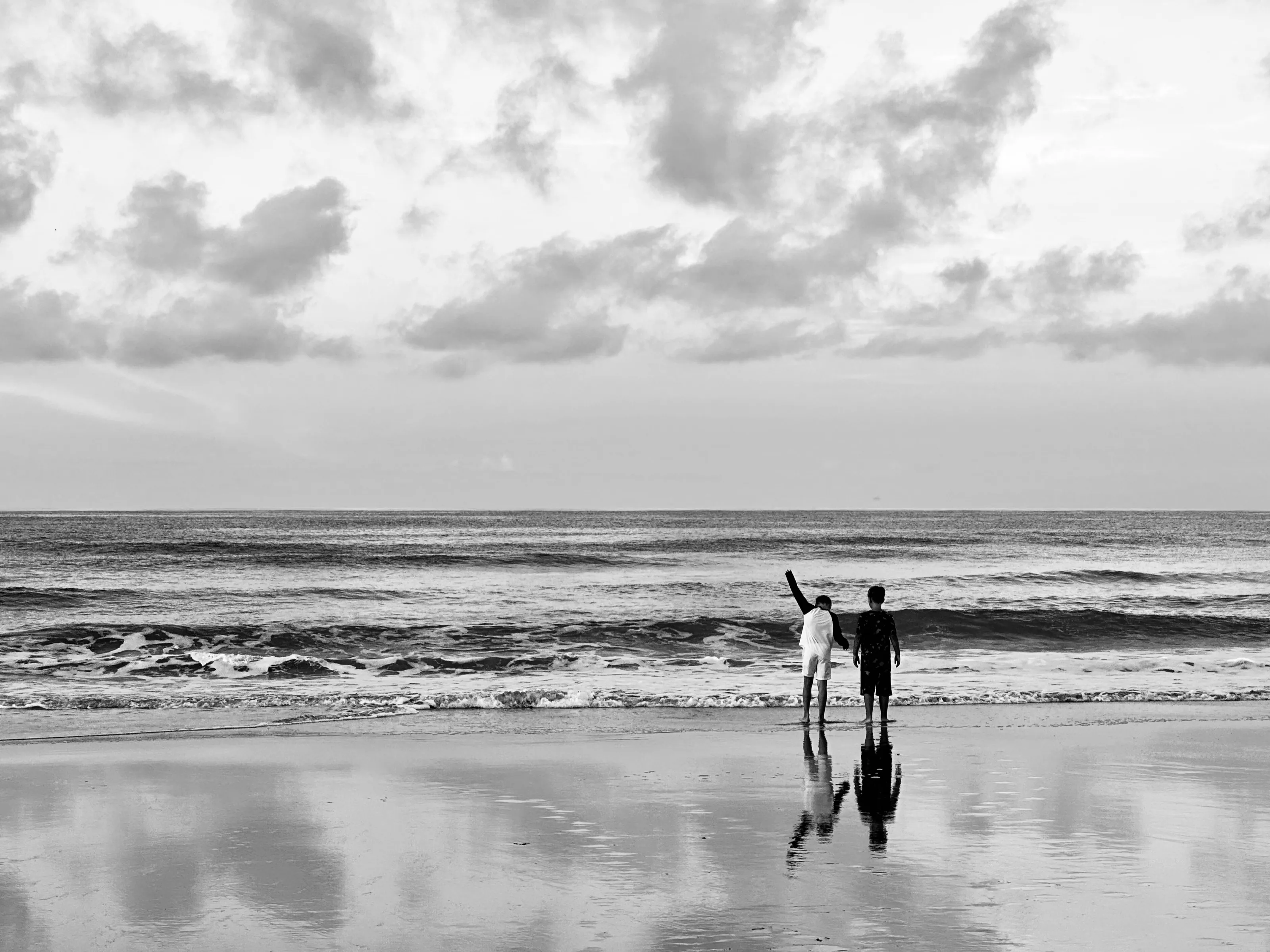 Two people stand on wet sand facing the ocean, reflected beneath a cloudy sky.