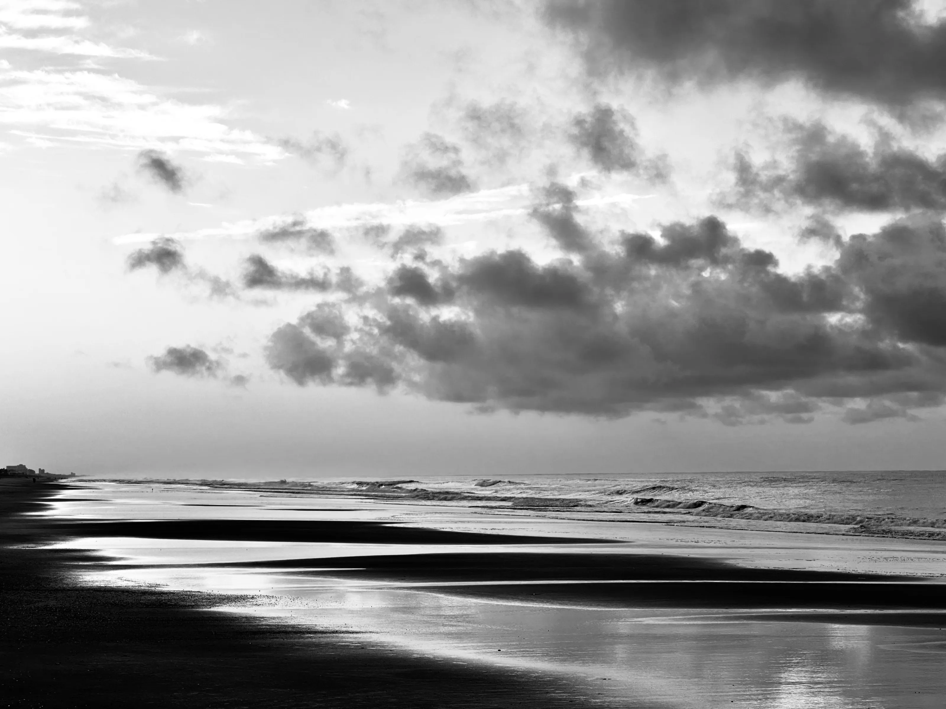Black-and-white beach scene with low waves, reflective wet sand, and large dark clouds overhead.