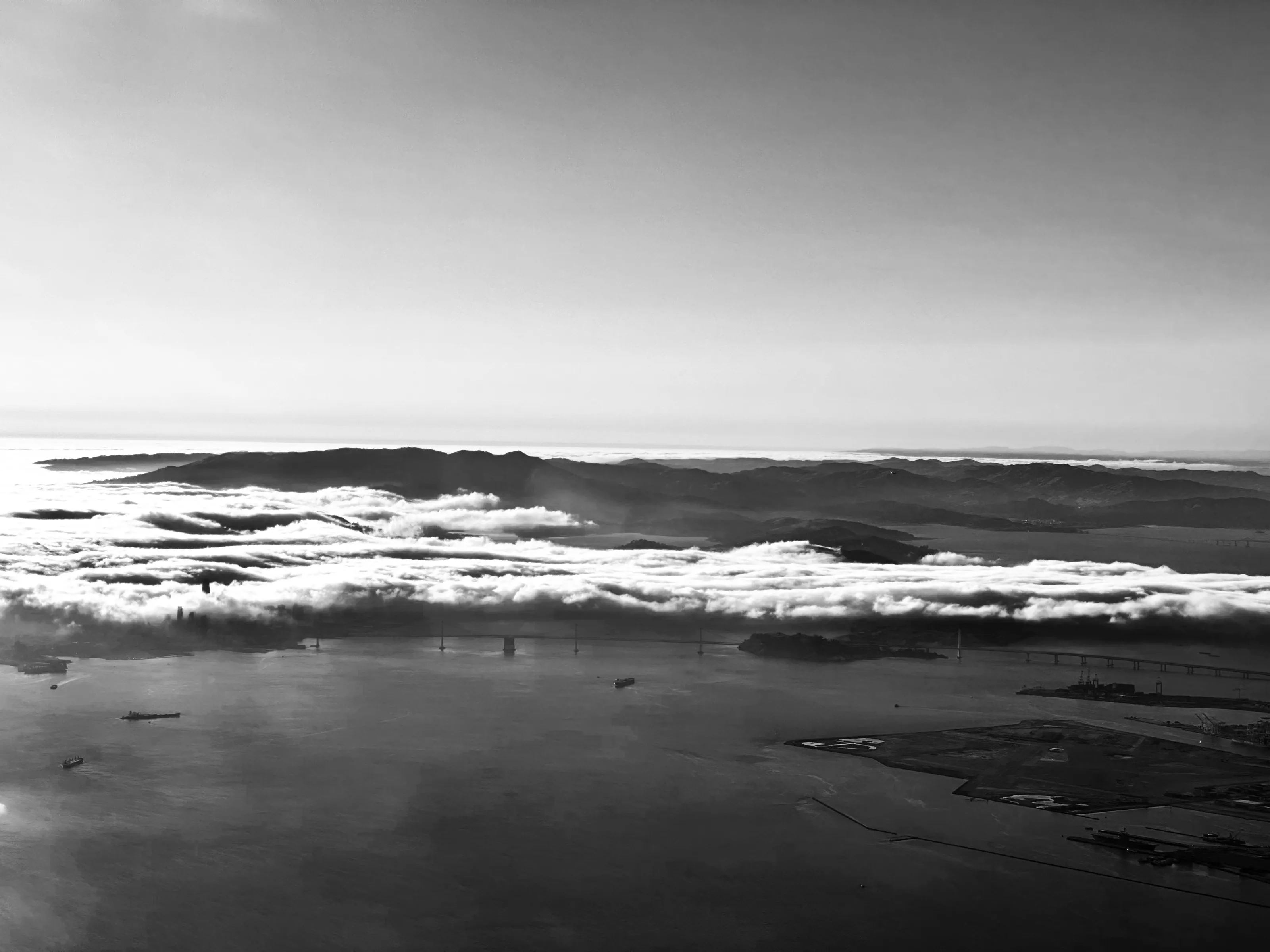 An aerial black-and-white view of a bay covered by a long band of fog, with hills on the horizon.